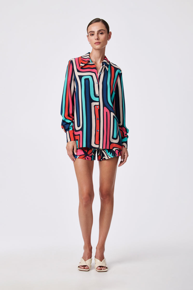 A woman poses against a white background wearing the Corbin 2 Short and a matching long-sleeve shirt, both featuring a bold geometric pattern in red, teal, black, white, and pink. She completes the look with white heeled sandals.