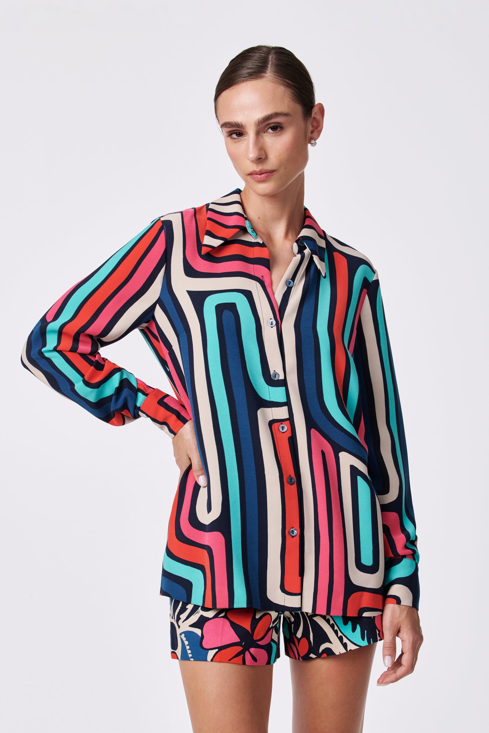 A woman stands against a plain white background with one hand on her hip, wearing the Montero Top in MULTI—a colorful, long-sleeve button-up shirt with bold geometric patterns—paired with patterned shorts.