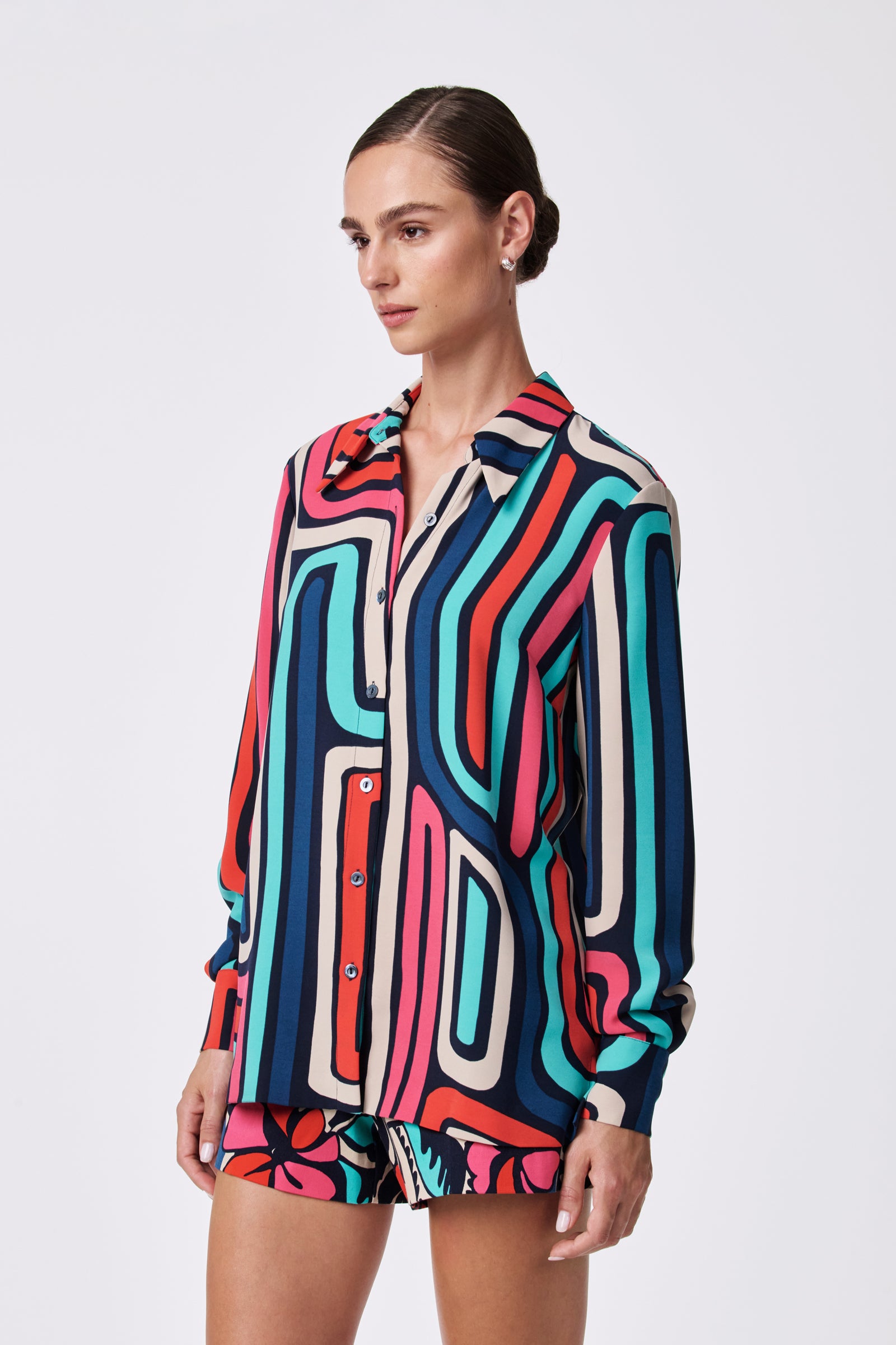 A woman poses against a plain background wearing the Montero Top, a colorful long-sleeve button-up shirt with bold abstract stripes in red, teal, navy, beige, and pink, paired with vibrant patterned shorts.