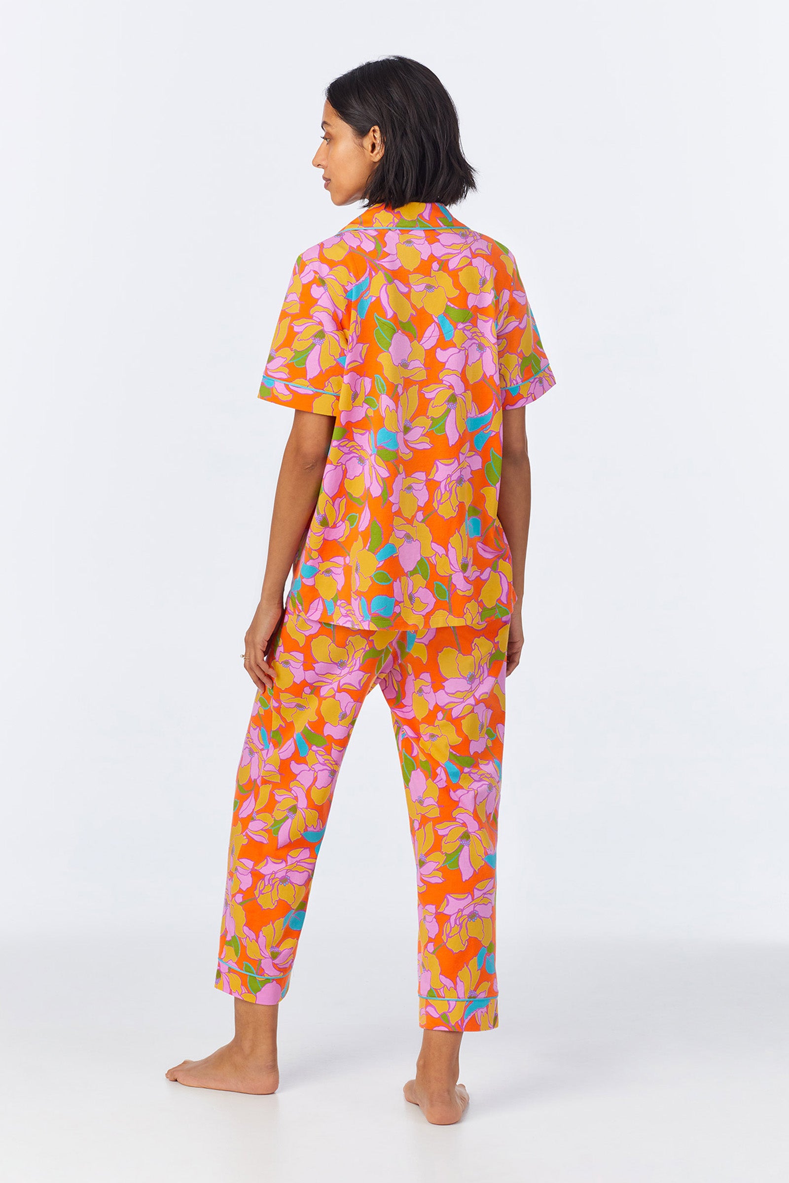 A person stands barefoot, facing away, wearing the Crop Classic PJ Set - Floral Poplin featuring a bold orange background with vibrant pink, purple, and yellow floral patterns.