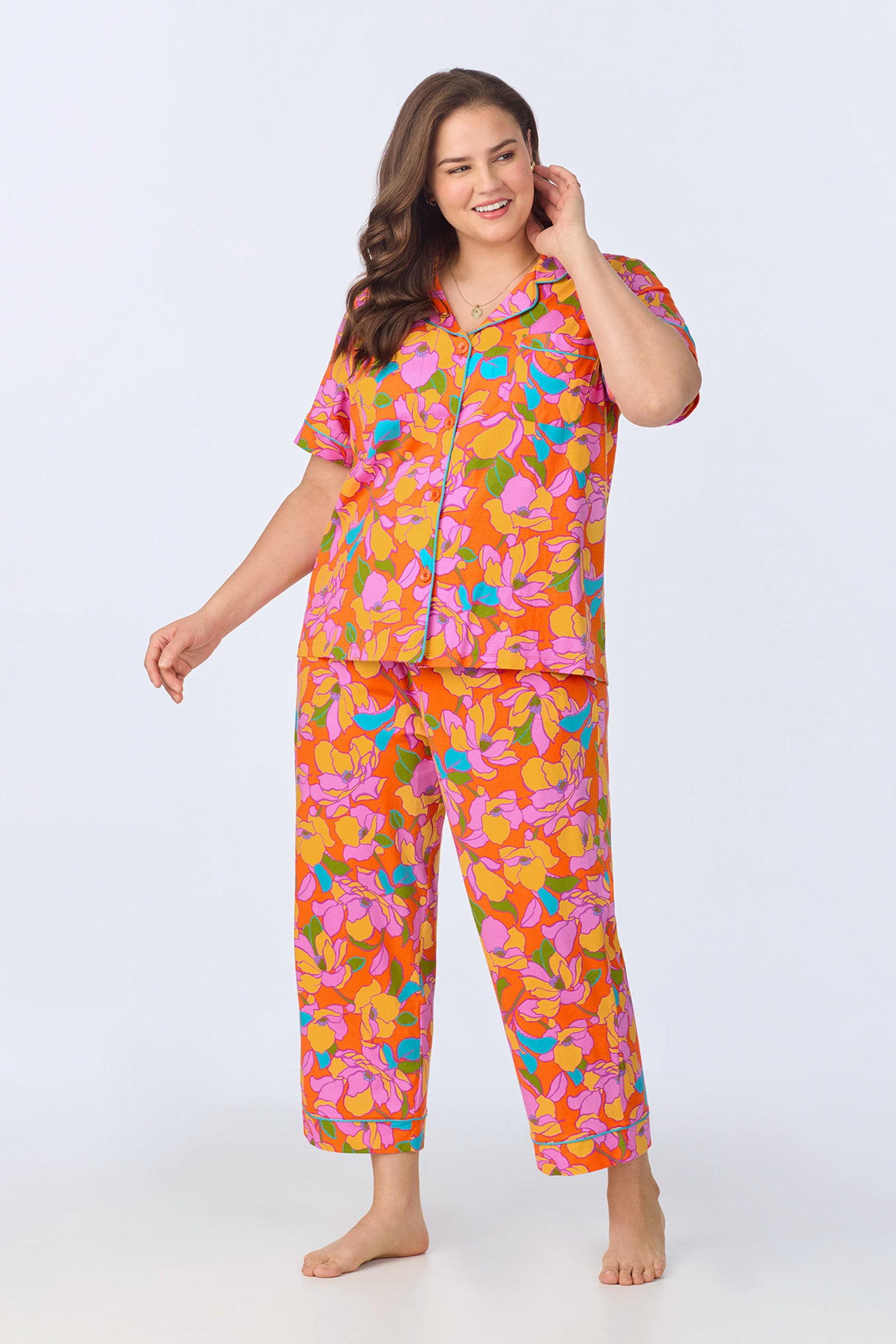 A woman smiles in the Crop Classic PJ Set - Floral Poplin, featuring a colorful floral pattern with a short-sleeve button-up top and matching pants. The background is plain and light-colored.
