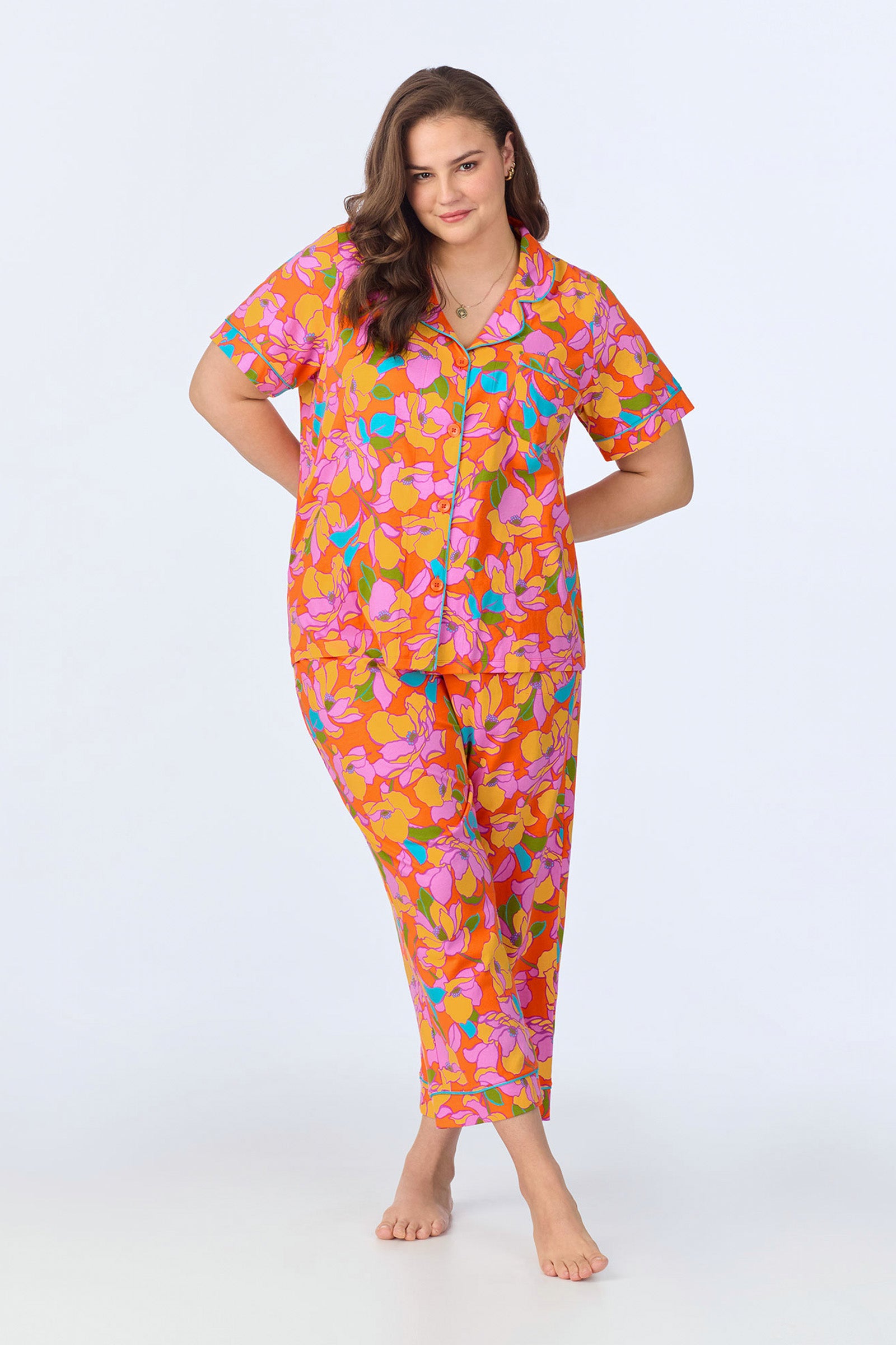 A woman poses barefoot in the Crop Classic PJ Set - Floral Poplin, a vibrant pajama set featuring a pink, orange, yellow, and blue floral pattern with a short-sleeve button-up top and matching pants against a plain background.