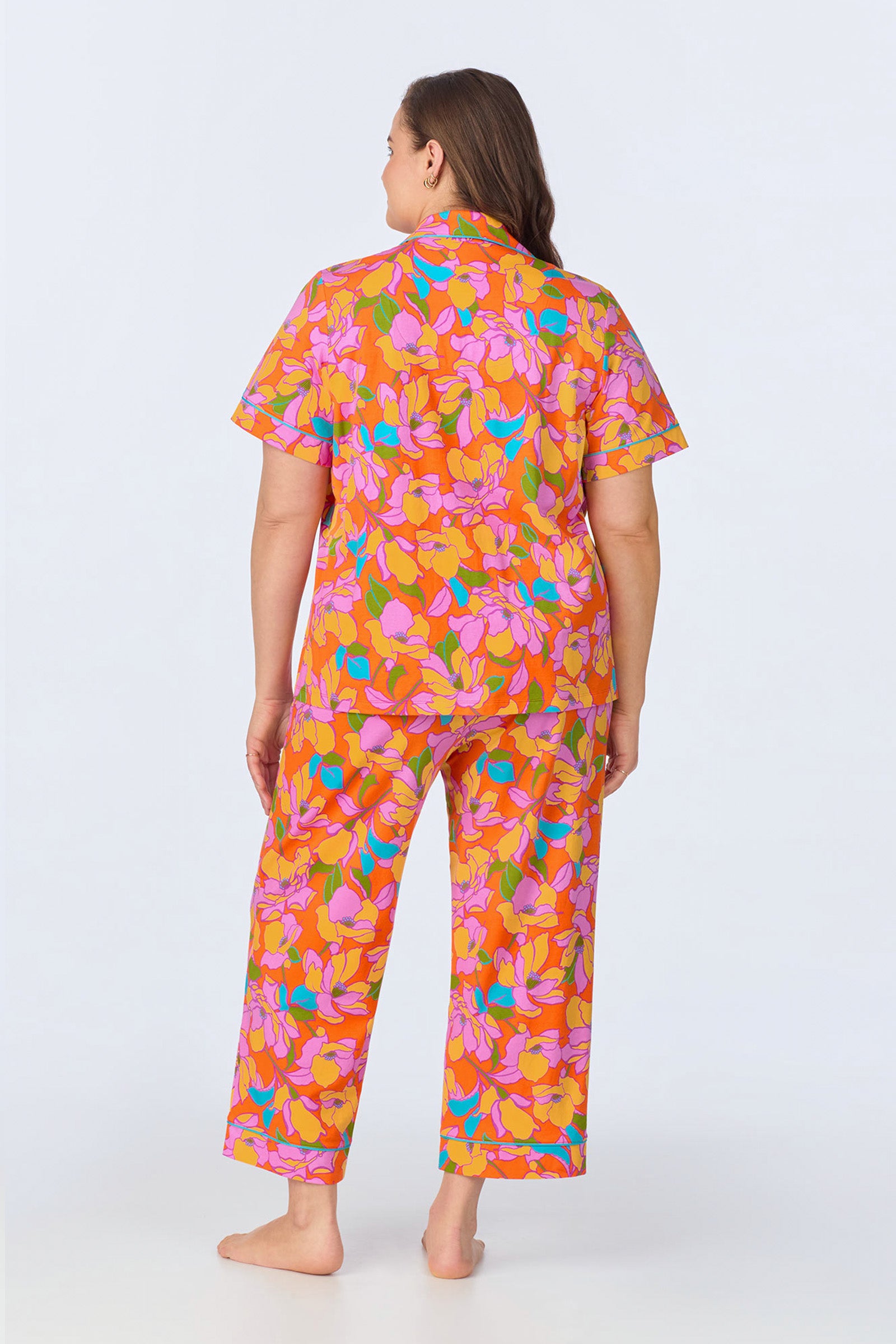 A woman stands barefoot facing away on a plain white background, wearing the Crop Classic PJ Set - Floral Poplin, featuring a colorful floral pattern in pink, orange, yellow, and blue with short sleeves and long pants.