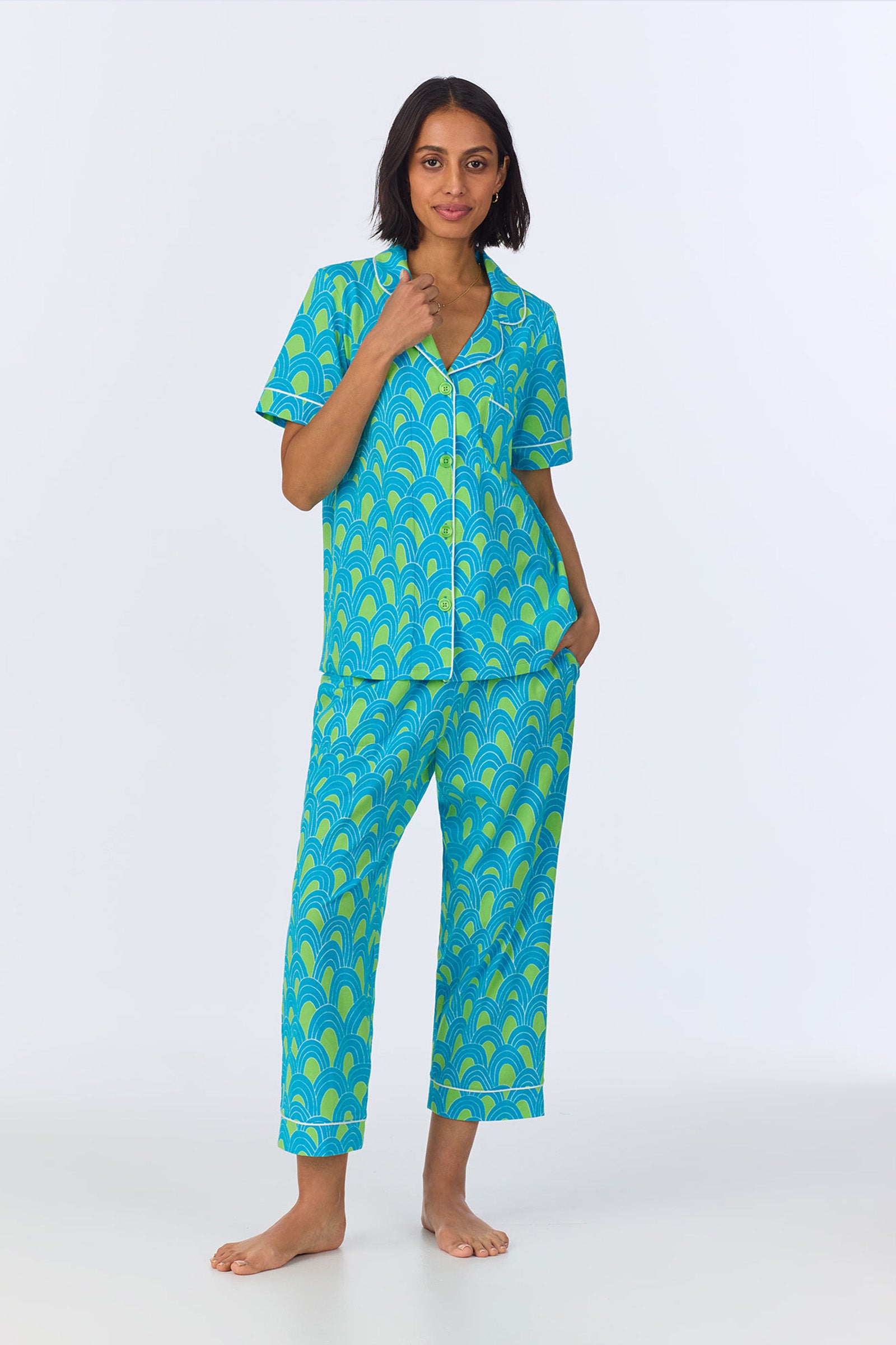 A woman stands barefoot in the Crop Classic PJ Set - Geo Stretch Jersey in Turquoise, featuring a matching short-sleeve button-up with a blue and green scallop pattern. She has one hand in her pocket and smiles slightly against a plain white background.