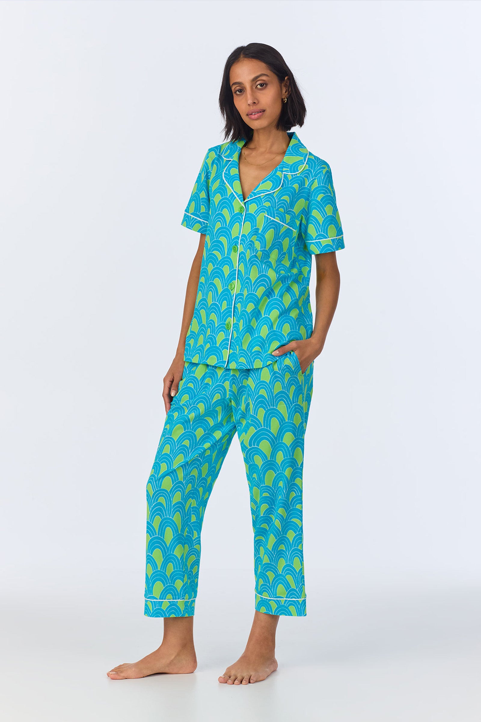 A woman stands barefoot in the Crop Classic PJ Set - Geo Stretch Jersey, featuring a blue and green scallop pattern, white piping and buttons, with one hand in her pocket against a plain light background.