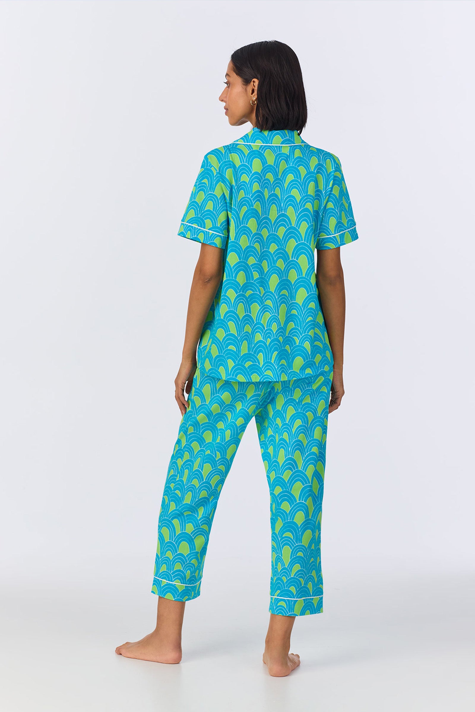 A woman stands barefoot facing away, wearing the Crop Classic PJ Set - Geo Stretch Jersey with short sleeves and ankle-length pants in a blue and green scallop pattern. The background is plain white.