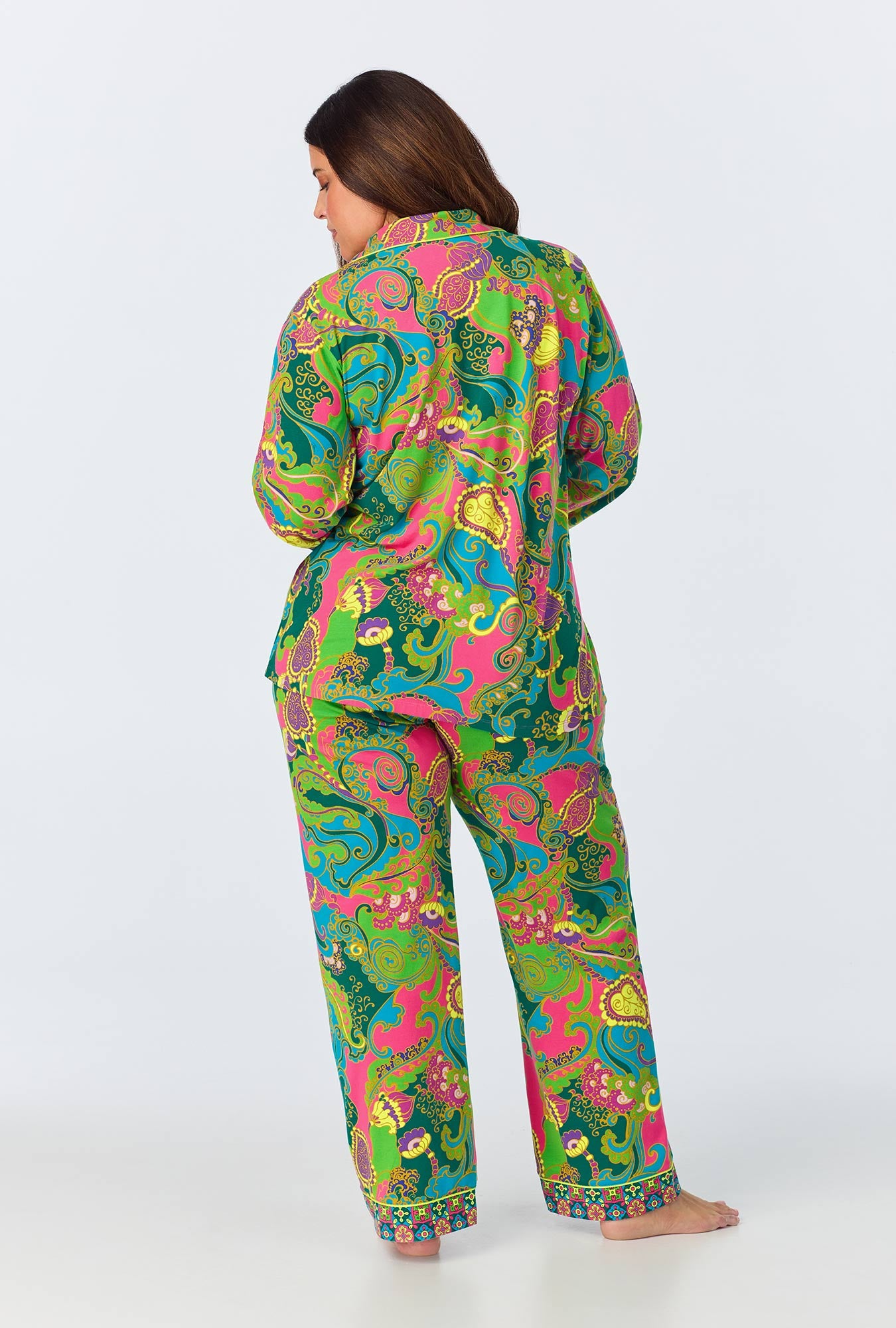A woman stands barefoot facing away, wearing the Turkish Delight Womens Long Sleeve Long Pant Jersey PJ Set with a bright, colorful paisley pattern in green, pink, yellow, and blue that pops against the plain white background.