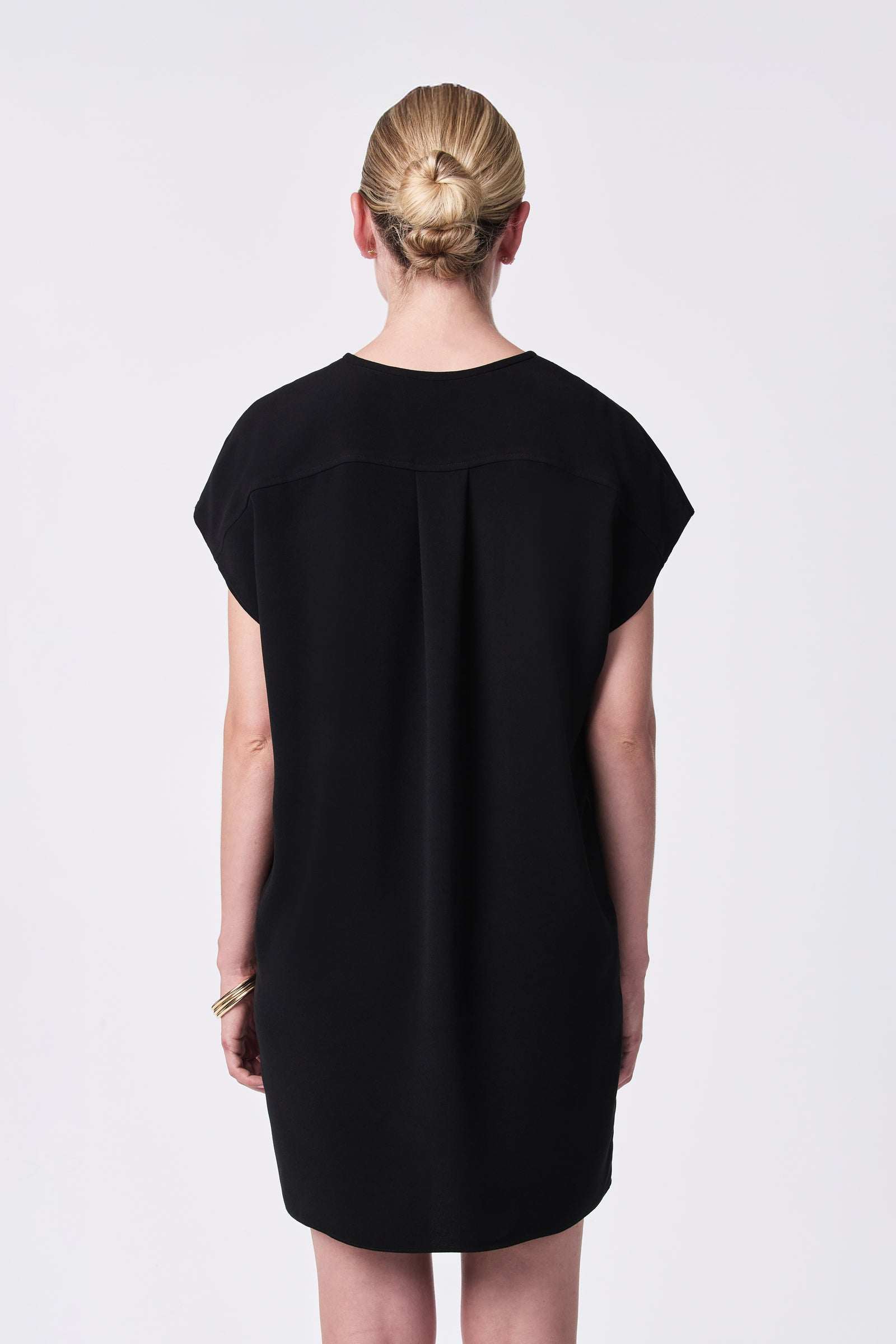 A woman with blonde hair in a bun wears the loose-fitting, short-sleeved black Hasil Dress. She stands facing away from the camera against a plain white background, accessorized with gold bangles on her left wrist.