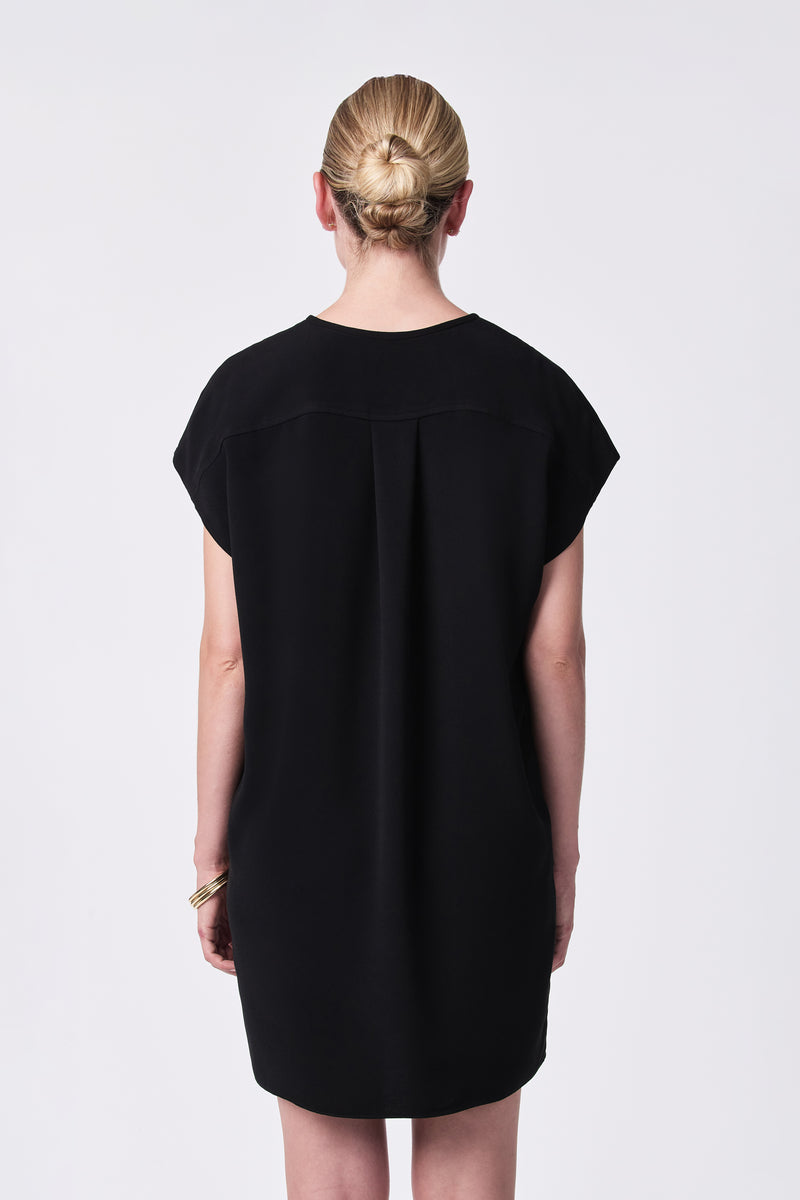 A woman with blonde hair in a bun wears the loose-fitting, short-sleeved black Hasil Dress. She stands facing away from the camera against a plain white background, accessorized with gold bangles on her left wrist.