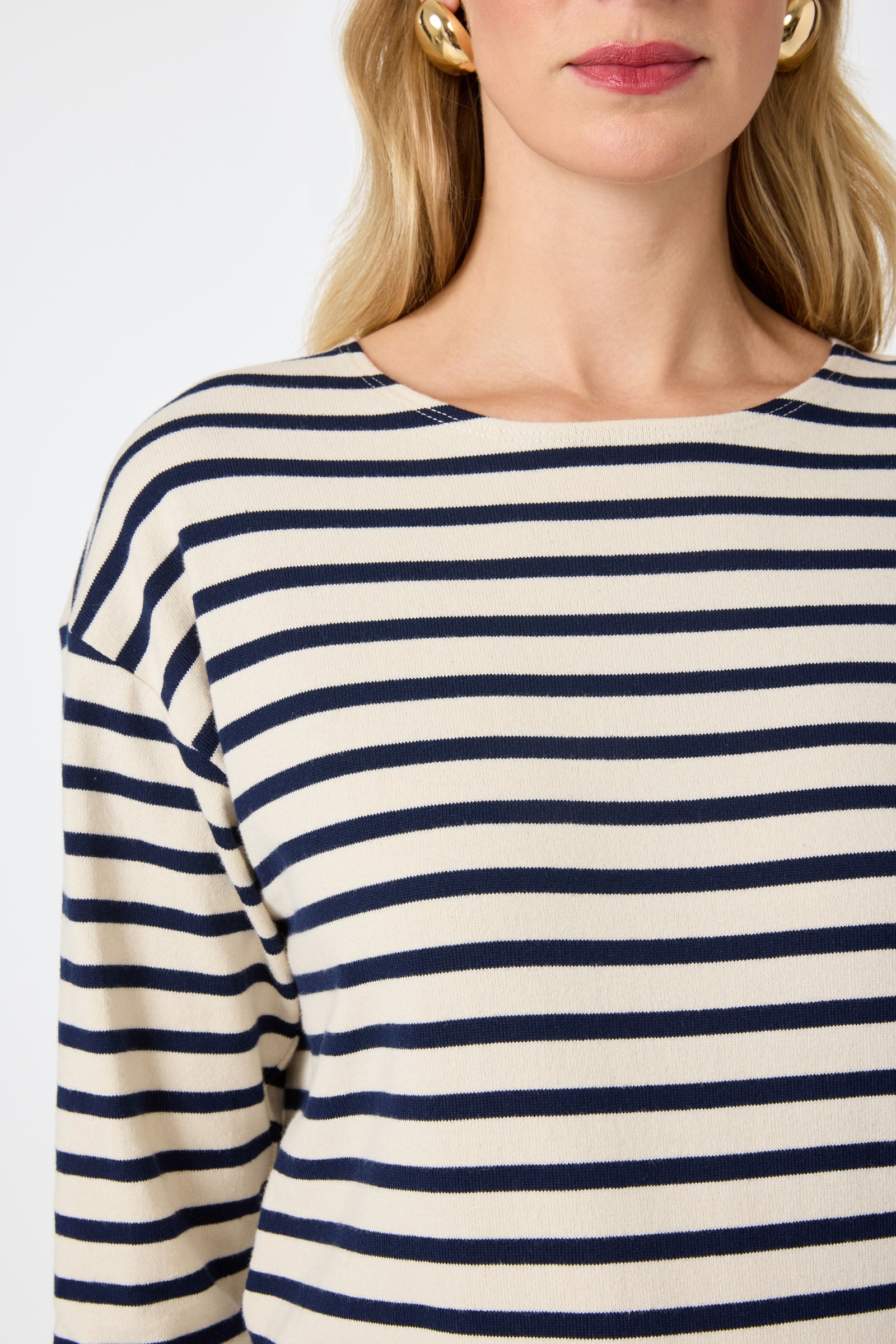 A woman wears the Novena Top in Noh Nightfall Stripe (cream and navy stripes) with gold hoop earrings. Her blond hair frames her partially visible face, highlighted by pink lipstick. Shown from shoulders to mid-chest.