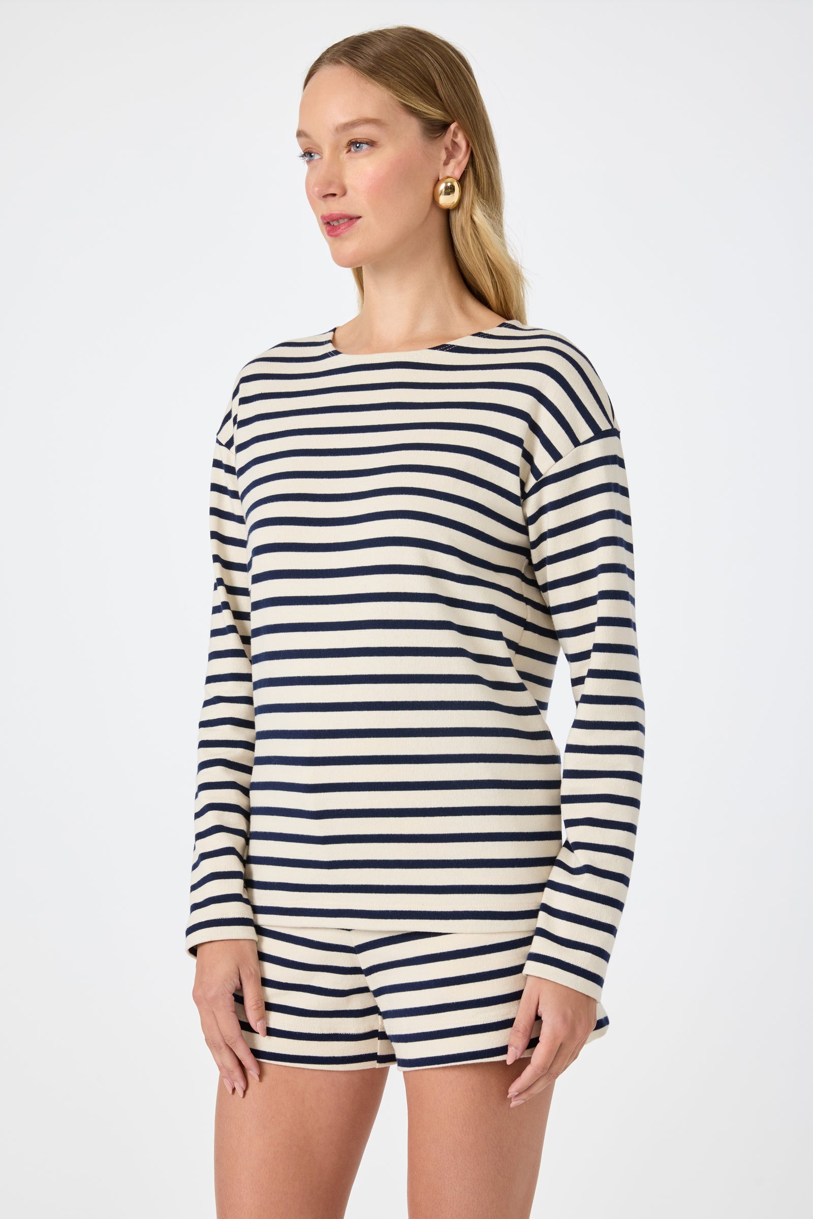 A woman wears the Novena Top in Noh Nightfall Stripe—long-sleeve, black and white stripes—paired with matching shorts, standing against a plain white background and gazing slightly to the side.