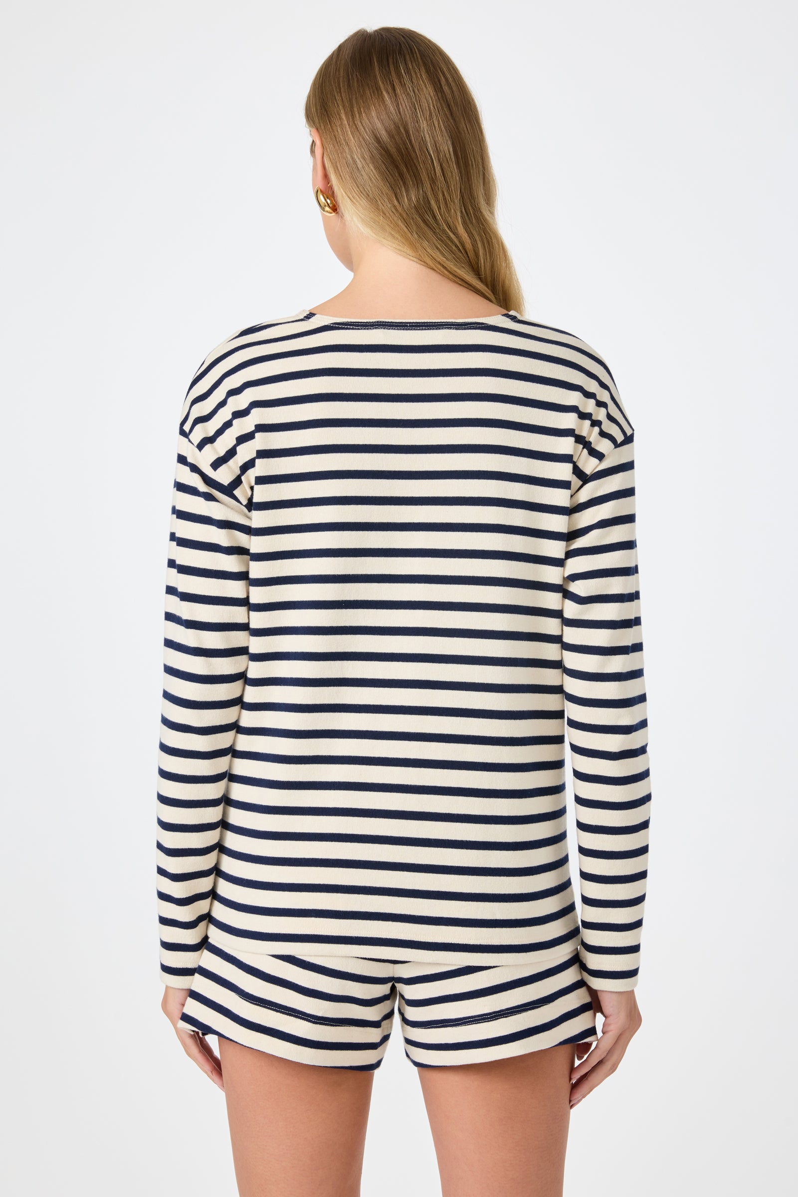 A woman with long, light brown hair is seen from behind wearing the Novena Top in Noh Nightfall Stripe, featuring cream and navy blue stripes, against a plain white background.