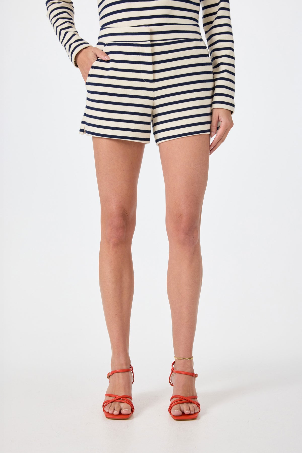 A person wearing Corbin 2 Shorts in Noh Nightfall Stripe with a matching long-sleeved top, red strappy sandals, and a gold anklet stands against a plain white background.