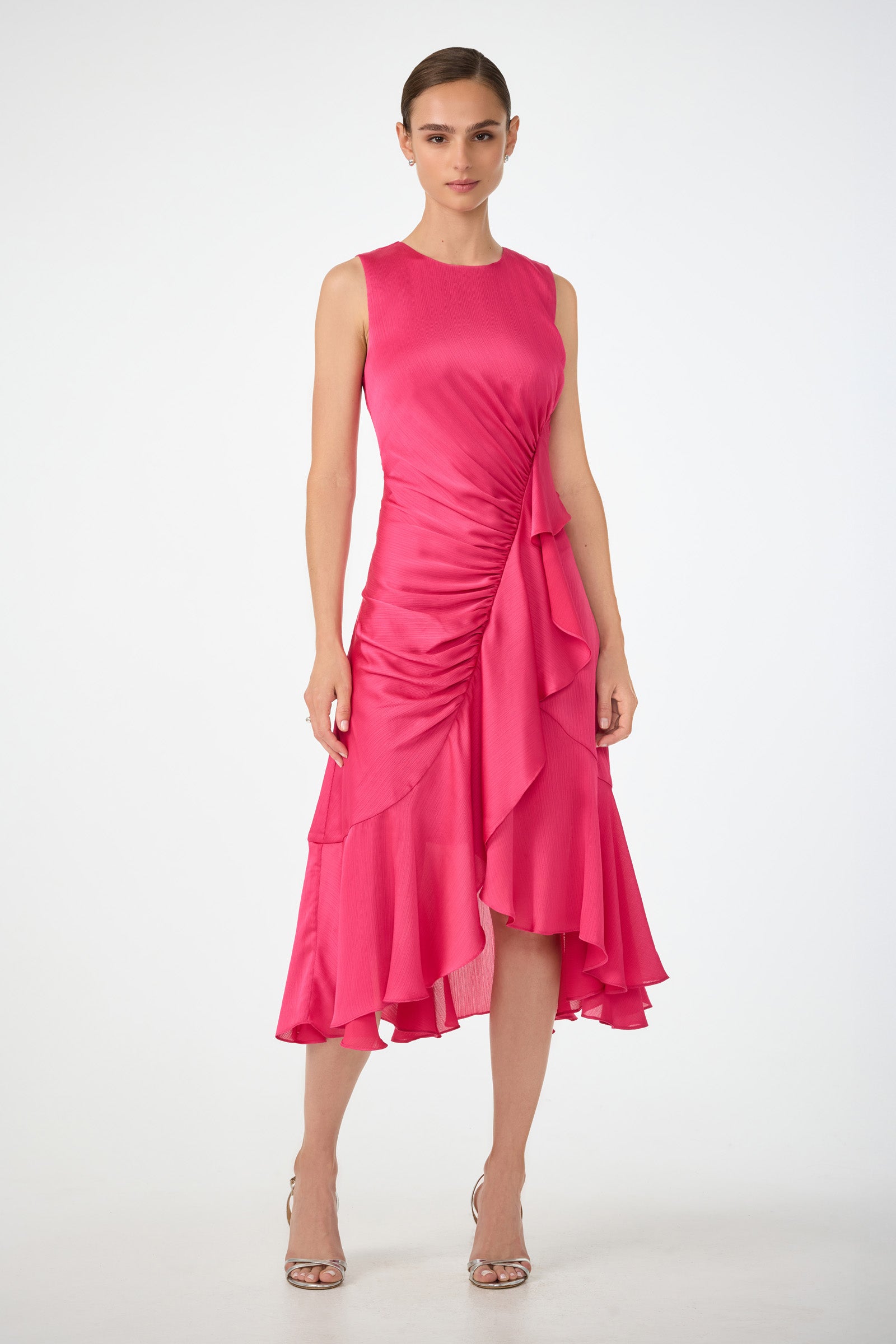 A woman poses in the Amara 2 Sleeveless Ruffle Dress in Forrest Rose, featuring an asymmetrical hem, ruffle details, and a gathered waist. She pairs it with open-toe heels and stands against a plain white background.