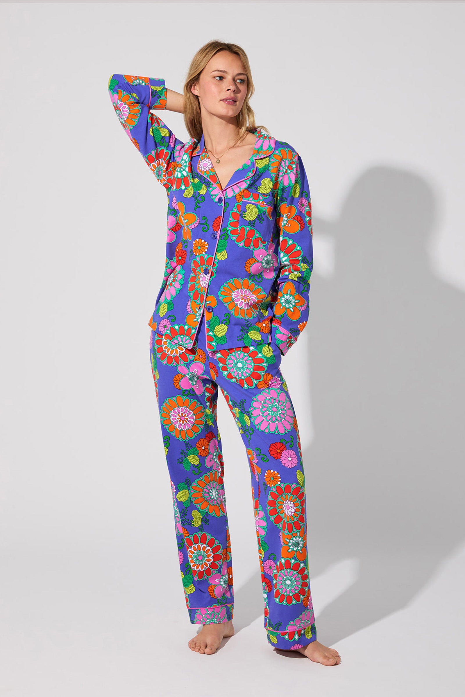 A woman stands barefoot against a light background, looking relaxed with one hand raised to her head, wearing the Classic PJ Set in the Tokyo Floral color, featuring a vibrant mix of blue, pink, orange, and green florals.