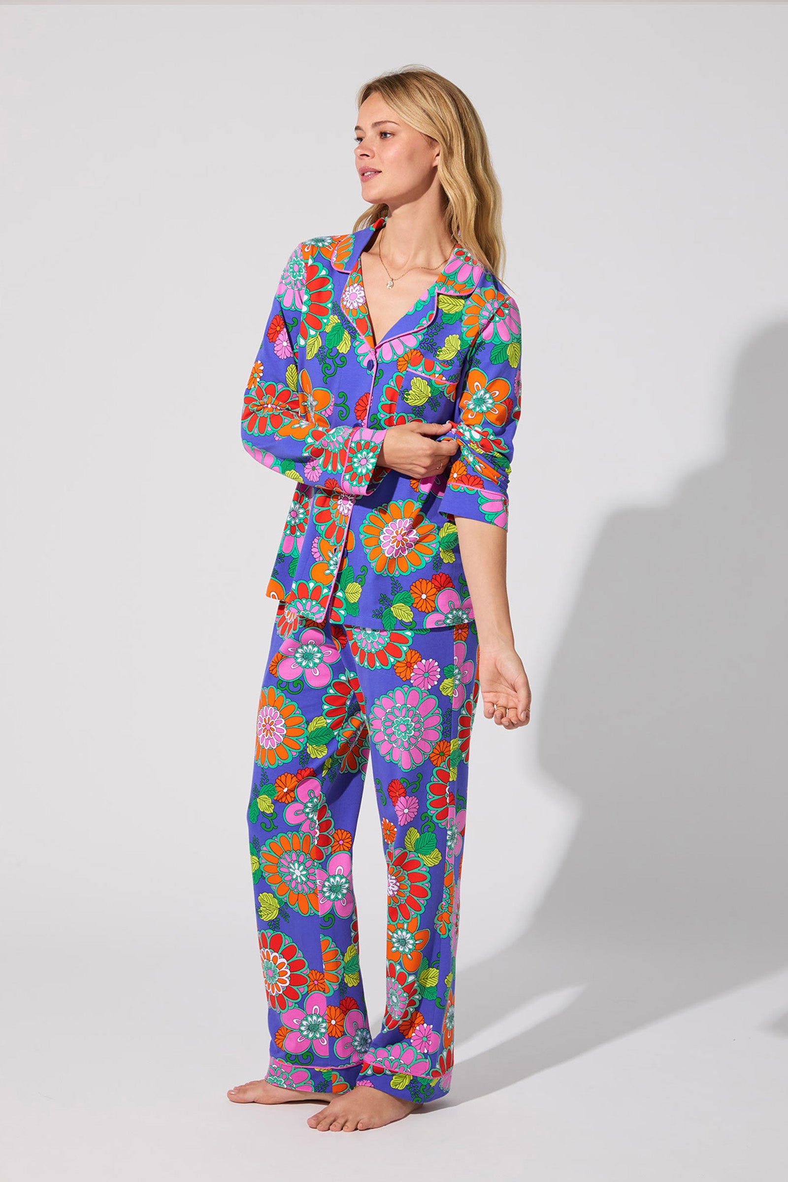 A woman stands barefoot in the Classic PJ Set - Tokyo Floral, featuring a bold floral pattern in purple, orange, red, pink, and green. She looks to the side with one hand raised against a plain light background.