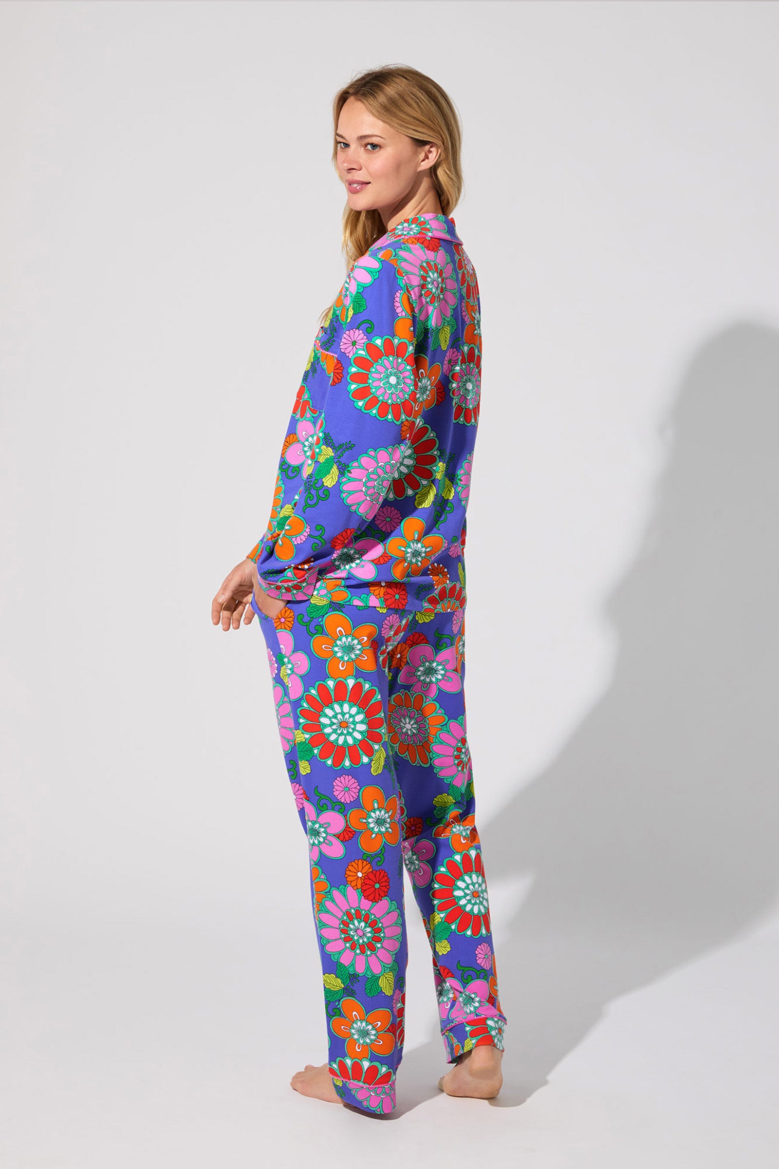 A woman stands barefoot against a plain background, smiling over her shoulder while wearing the Classic PJ Set - Tokyo Floral, a colorful loose-fitting pajama set with a bold red, blue, green, and orange floral pattern.