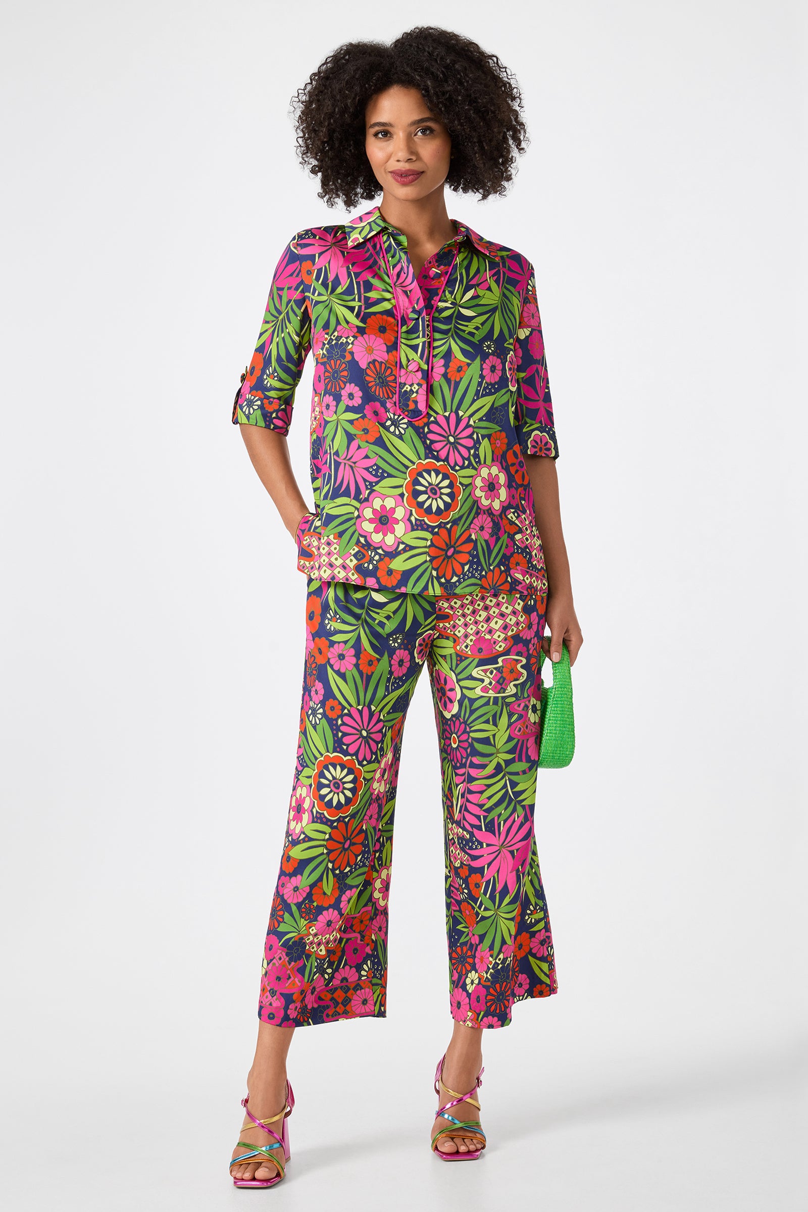 A woman stands confidently in the Estella 2 Cropped Pants - Fumi Floral, paired with a matching floral shirt in vibrant pink, green, and orange. She accessorizes with a small green purse and platform sandals.