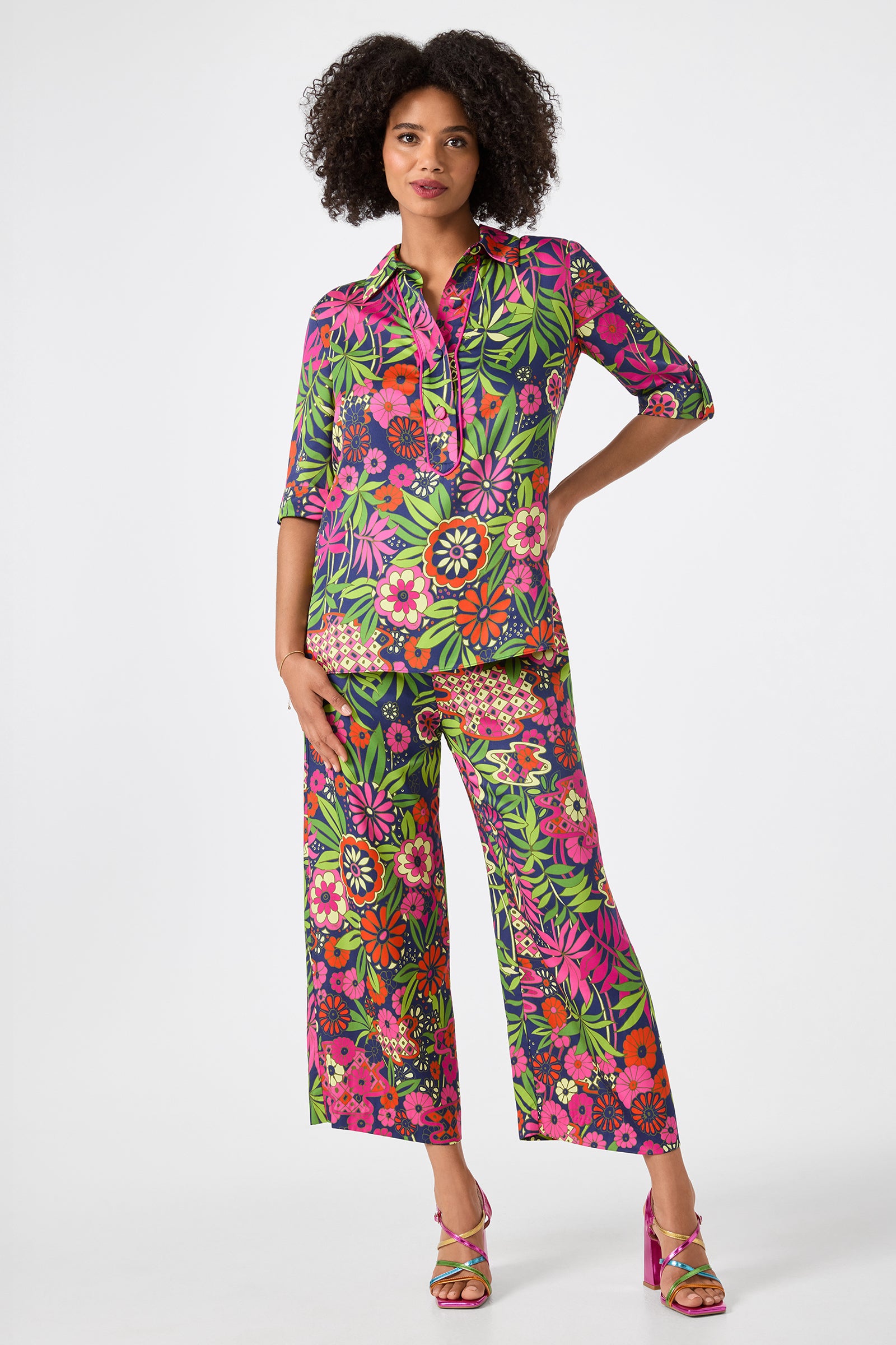 A woman poses against a white background wearing the Portrait 2 Top - Fumi Floral, a vibrant, floral-patterned button-up shirt paired with wide-leg pants in pink, green, yellow, and orange tones and pink high-heeled sandals.
