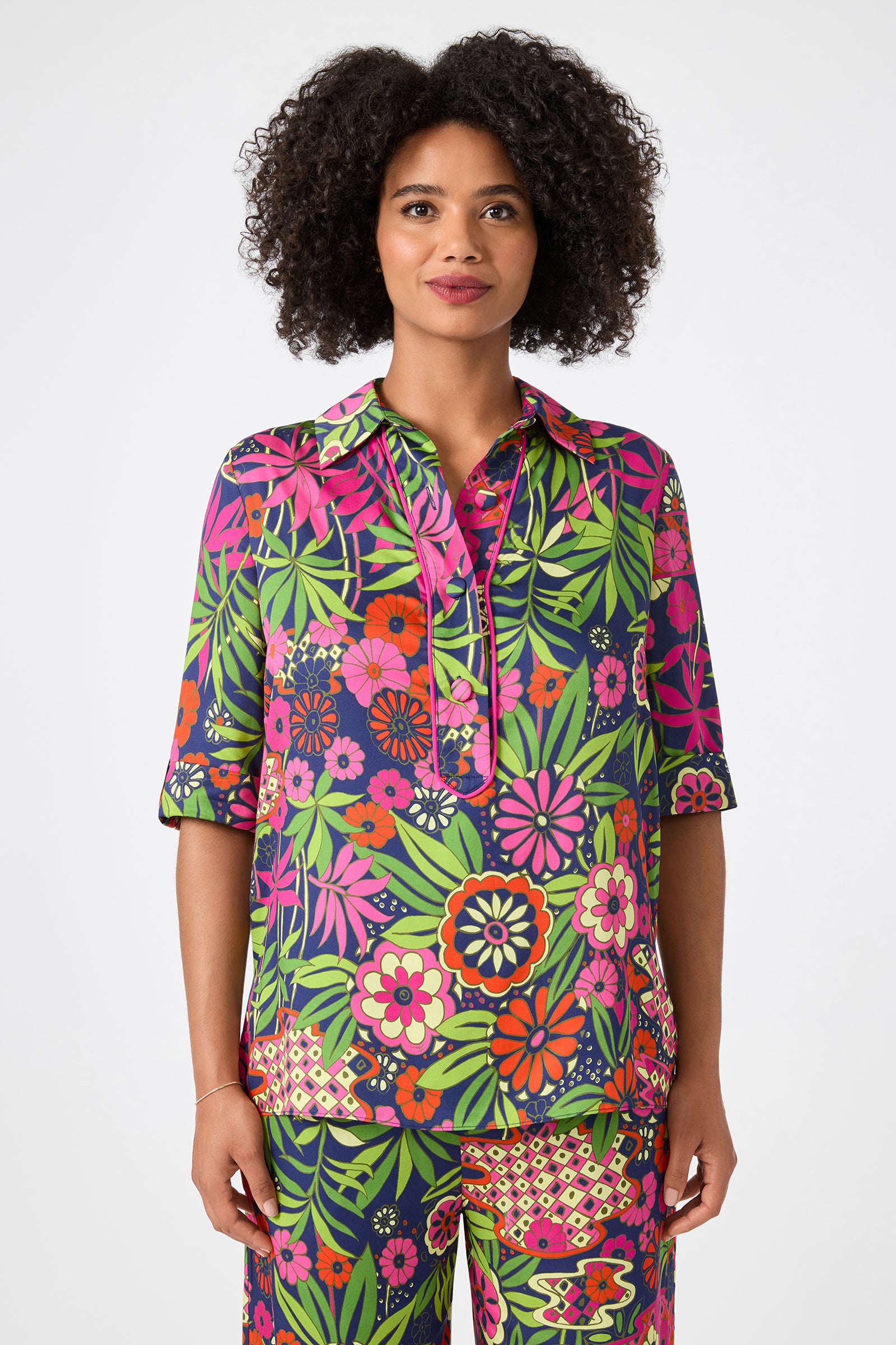 A woman with curly hair wears the Portrait 2 Top - Fumi Floral, a colorful shirt featuring vibrant green, pink, purple, red, and orange floral patterns, standing against a plain white background.