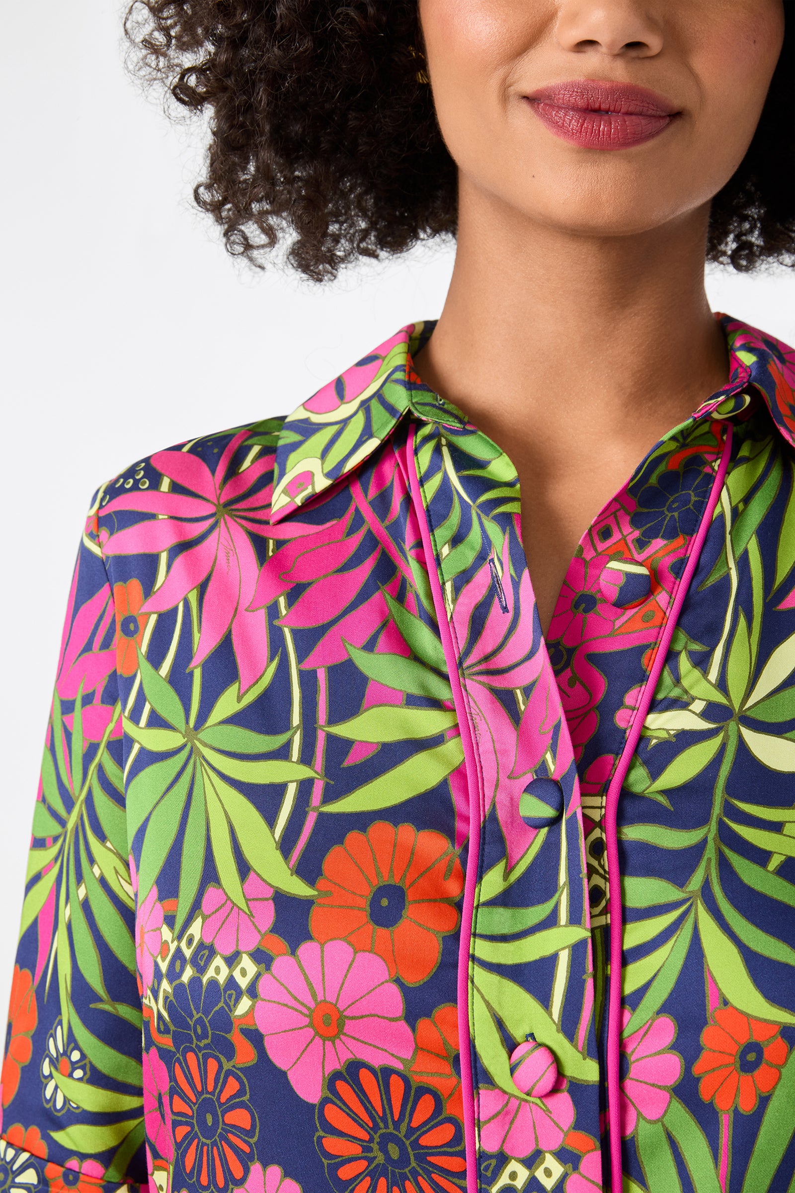 A woman with curly hair wears the Portrait 2 Top - Fumi Floral, featuring bright pink, green, and orange flowers and leaves. She stands against a plain white background, with only the lower half of her face visible.