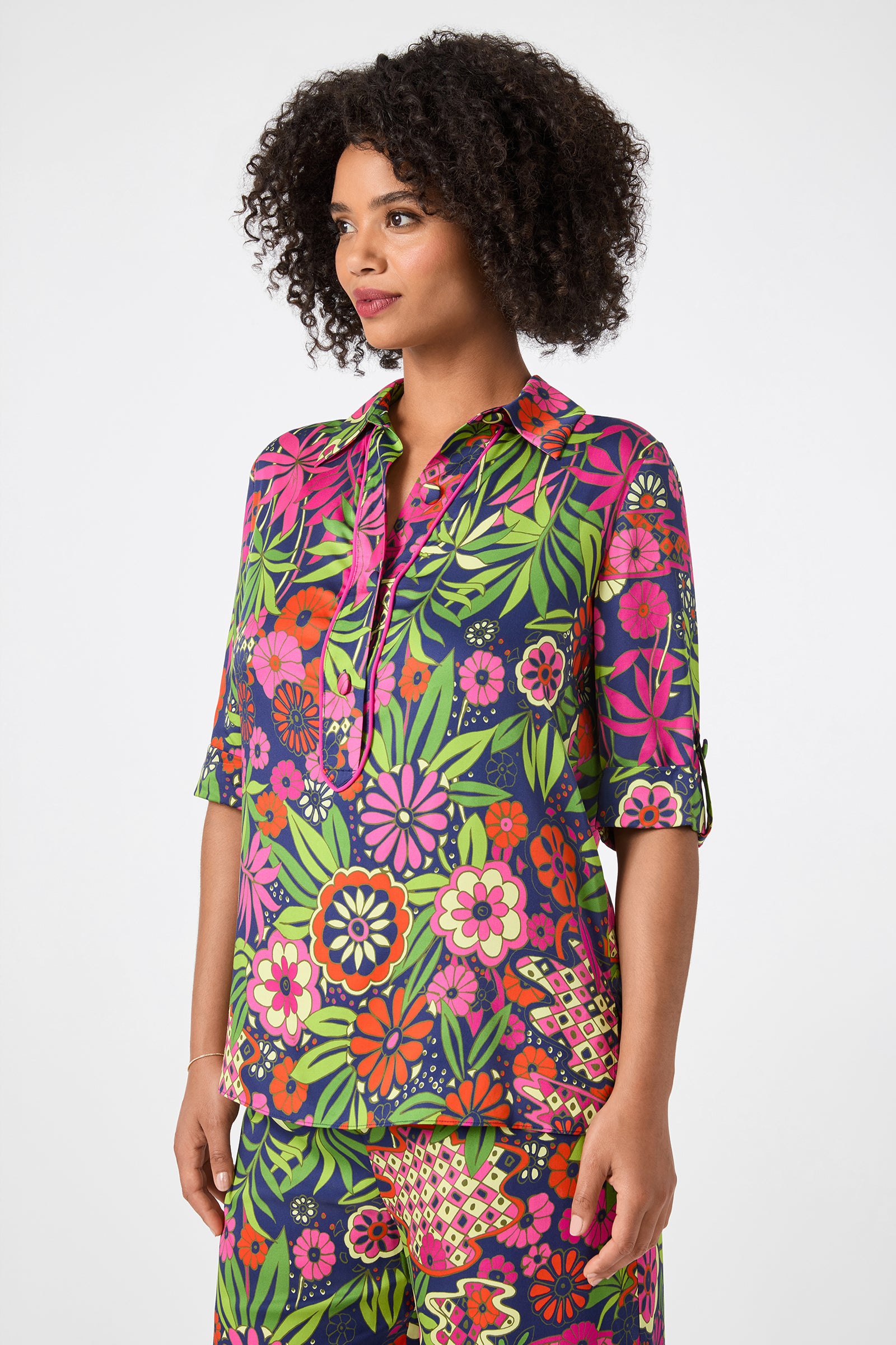 A woman with curly hair wears the Portrait 2 Top - Fumi Floral, a colorful blouse featuring vibrant pink, green, orange, and yellow floral and leaf patterns. She stands against a plain white background.