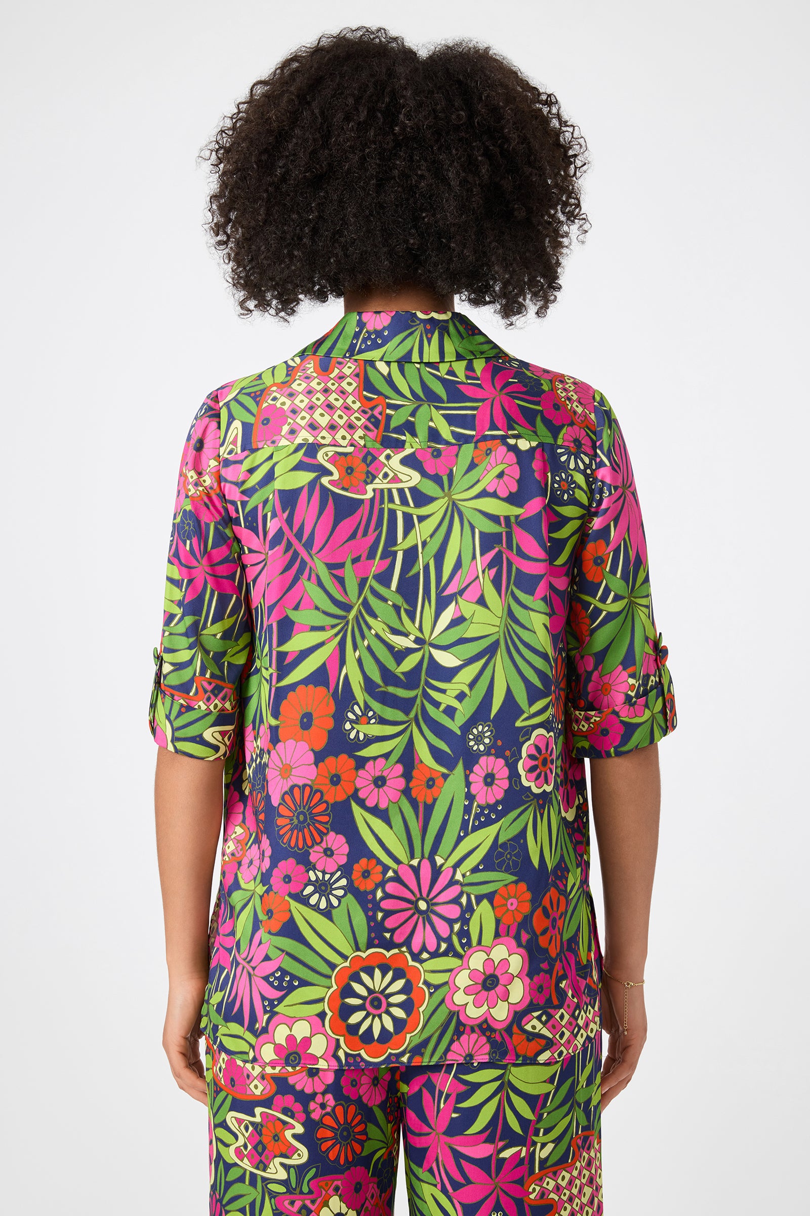 A person with medium-brown skin and curly hair faces away, wearing the Portrait 2 Top - Fumi Floral, featuring a bold green, pink, red, and orange floral pattern. The background is plain white.