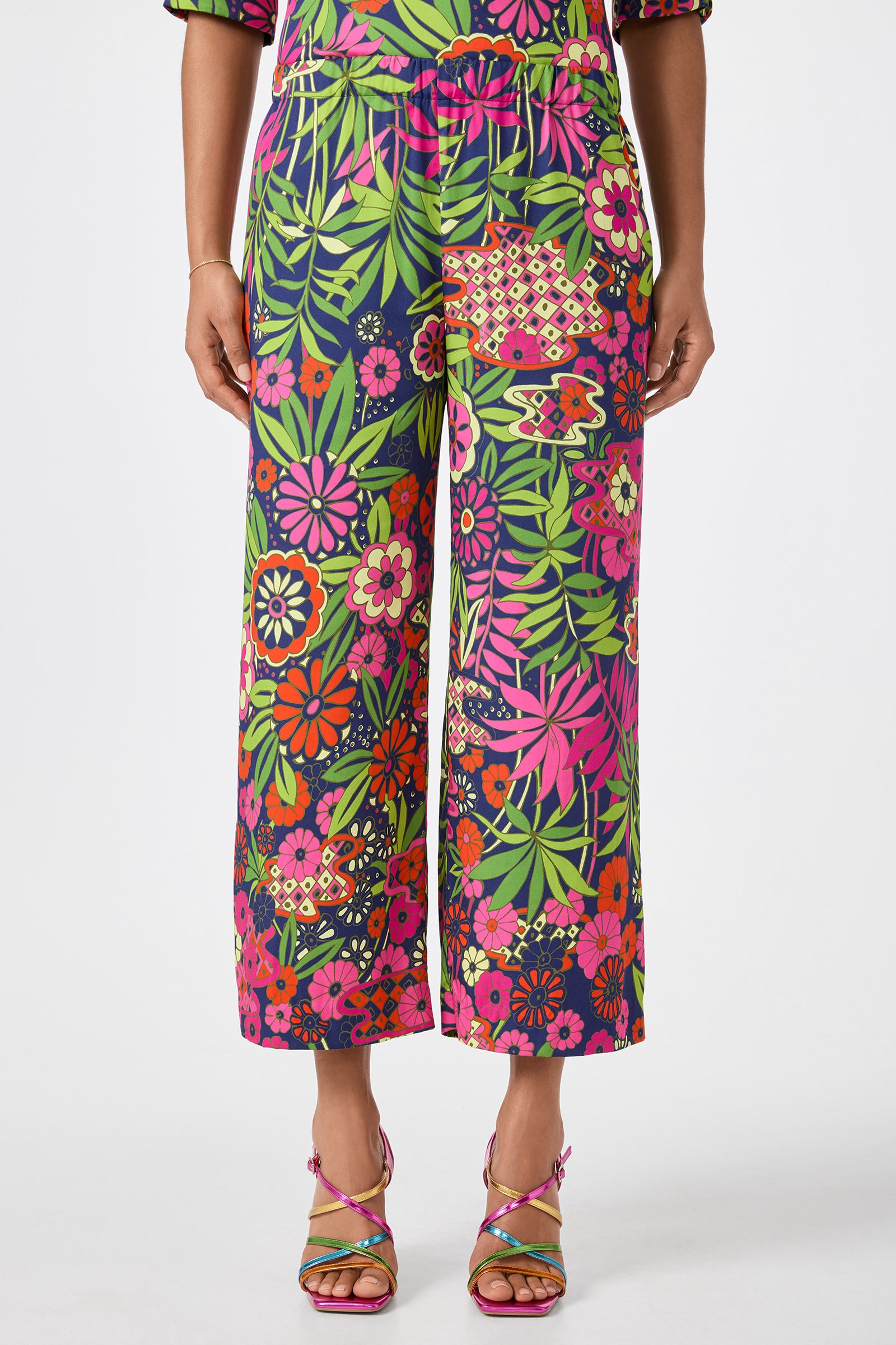 A person stands against a plain white background wearing Estella 2 Cropped Pants in Fumi Floral—a high-waisted, wide-leg style with a vibrant pink, red, green, and white floral pattern—paired with colorful strappy sandals.