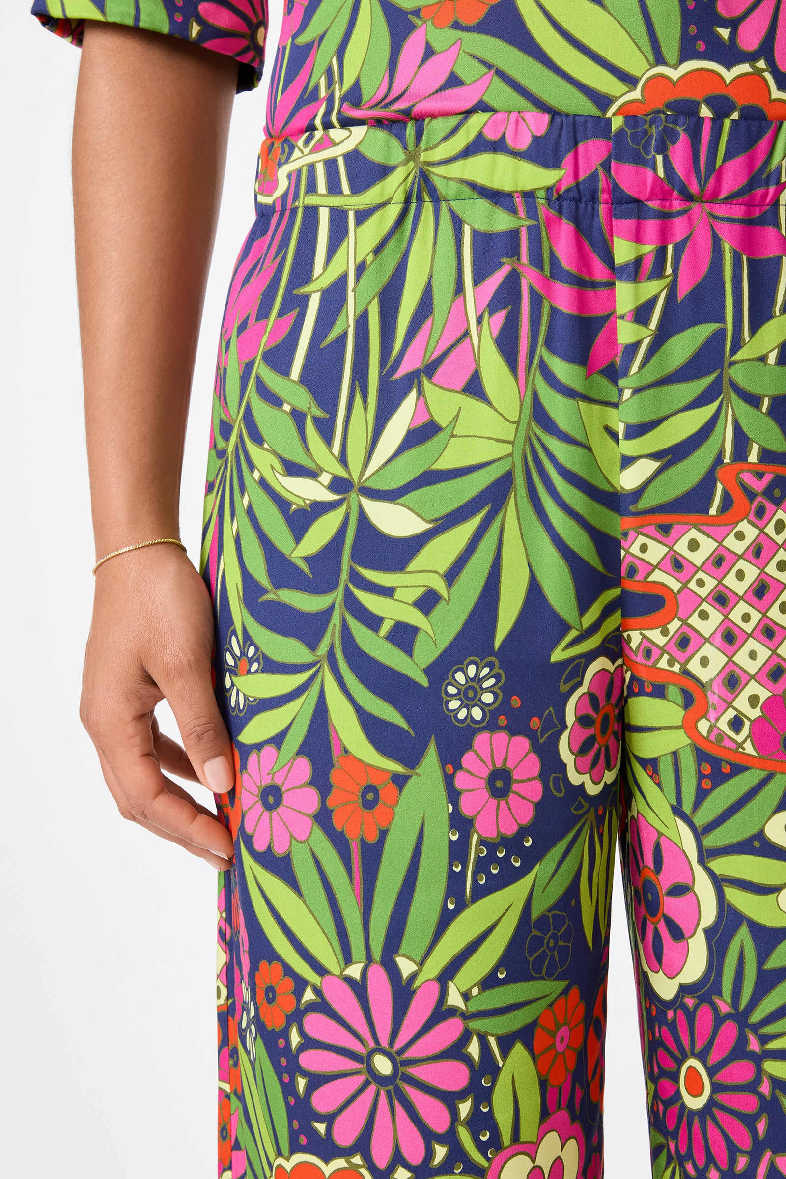A person wearing Estella 2 Cropped Pants - Fumi Floral, featuring a bold green, pink, red, and blue floral and leaf pattern. Only the lower torso and upper legs are visible, with their left hand resting by their side.