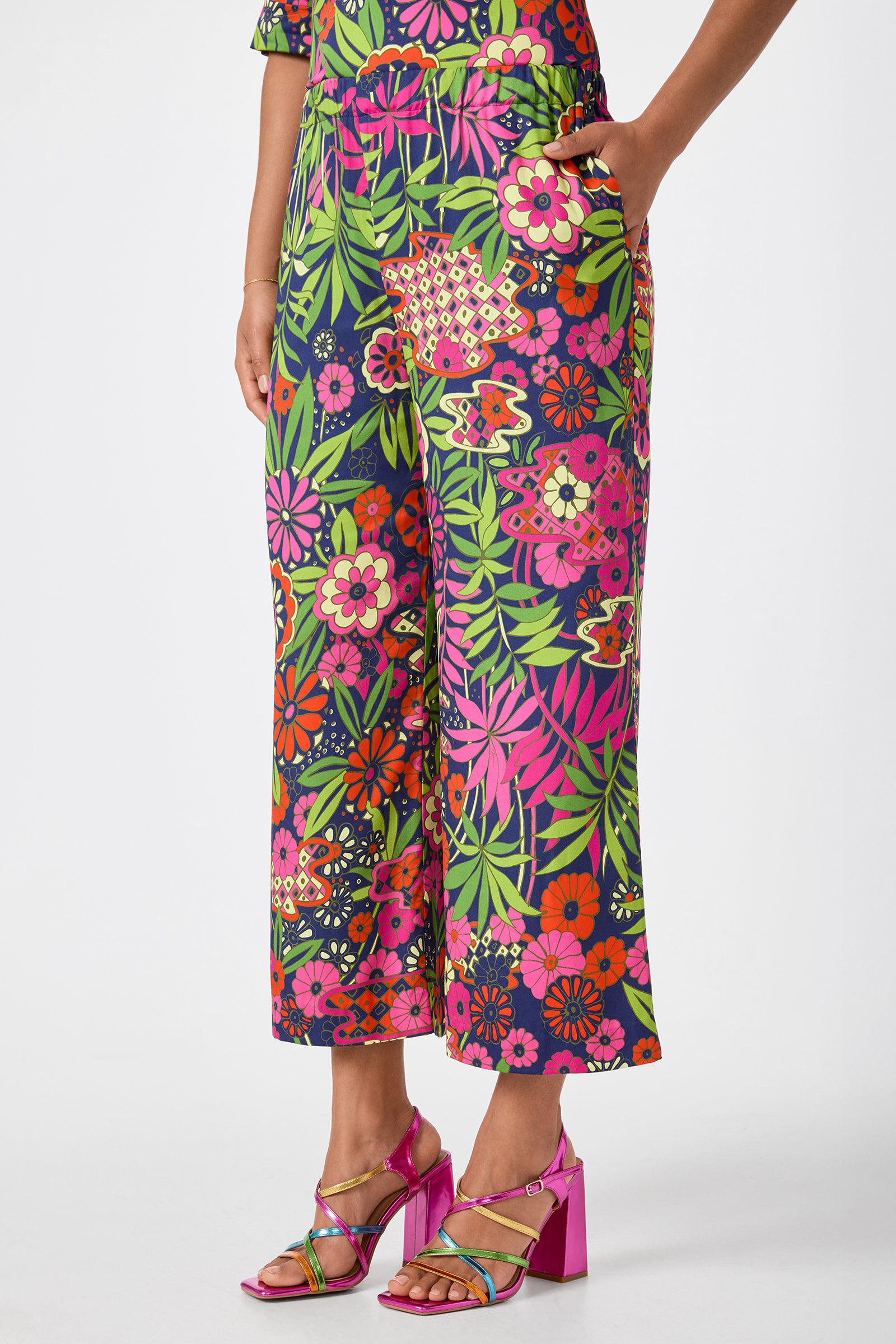 A person wearing Estella 2 Cropped Pants - Fumi Floral, which feature a vibrant floral pattern in pink, green, orange, and yellow, paired with shiny pink high-heeled sandals against a plain white background.