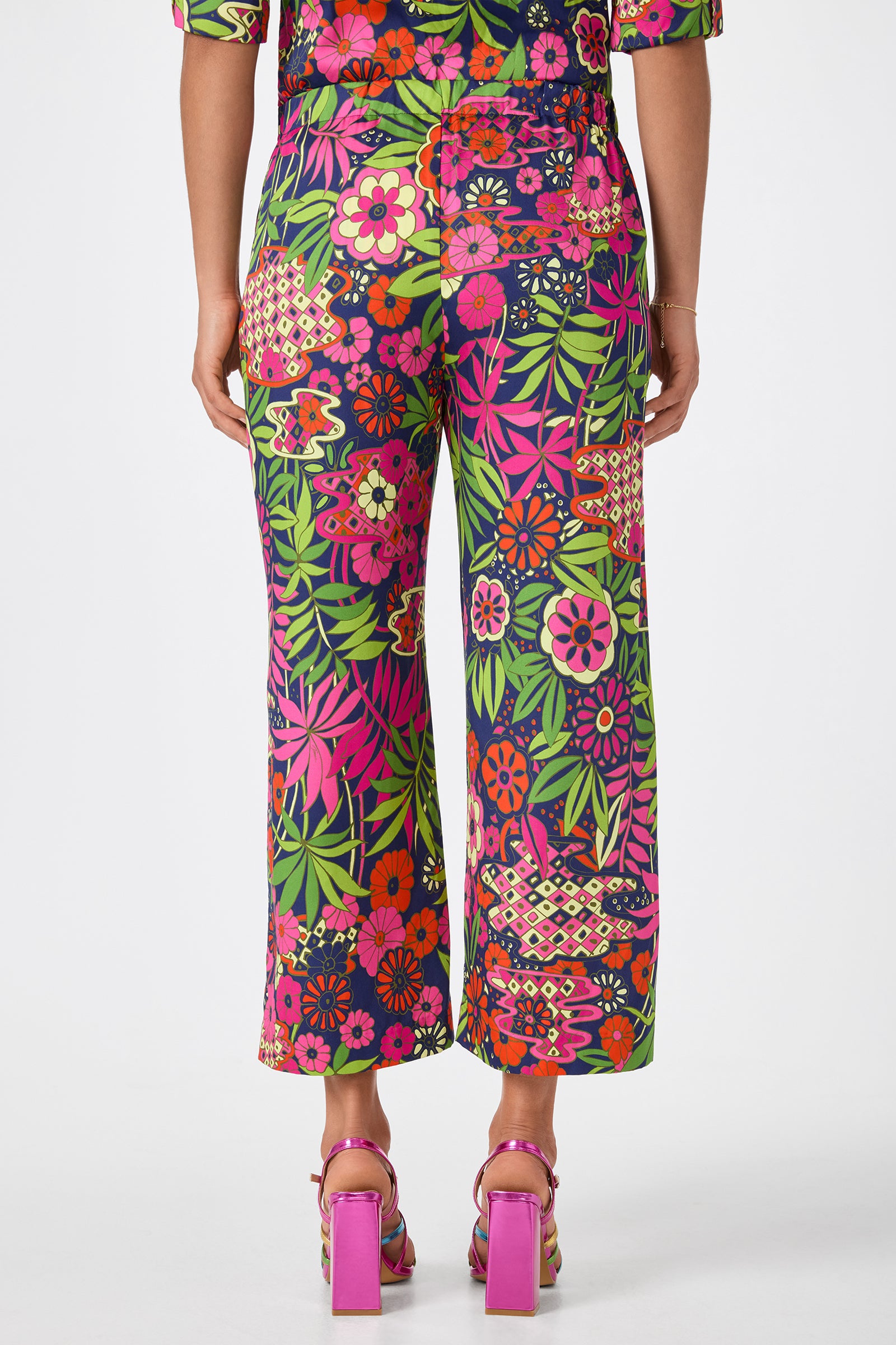 A person, seen from behind and cropped at the upper back, wears Estella 2 Cropped Pants – Fumi Floral in vibrant pink, orange, green, and purple prints with pink block-heeled shoes.