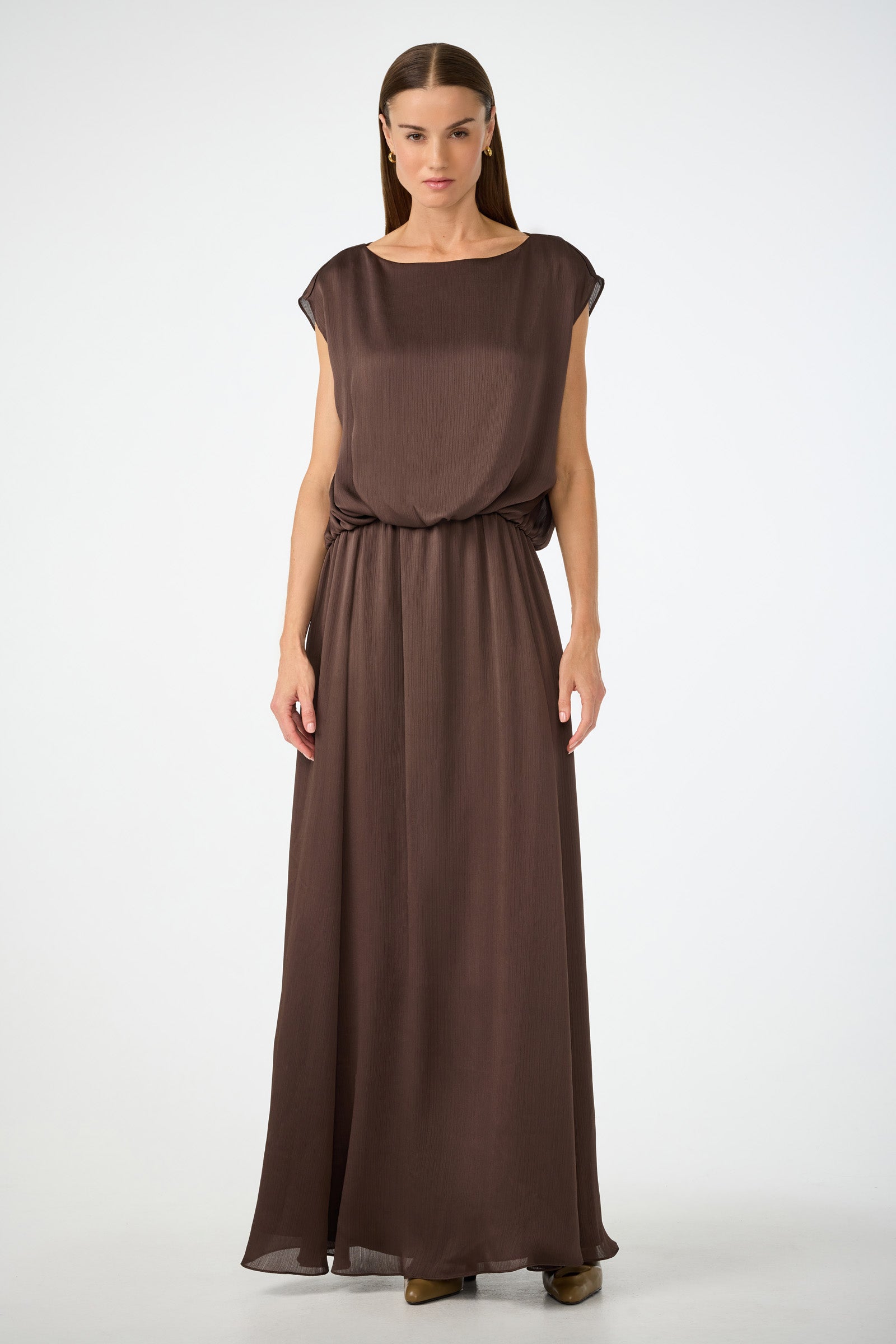 A woman with straight brown hair and a neutral expression wears the Vantage 2 Maxi Dress—a sleeveless, floor-length brown dress with a gathered waist and loose fit—standing against a plain white background.