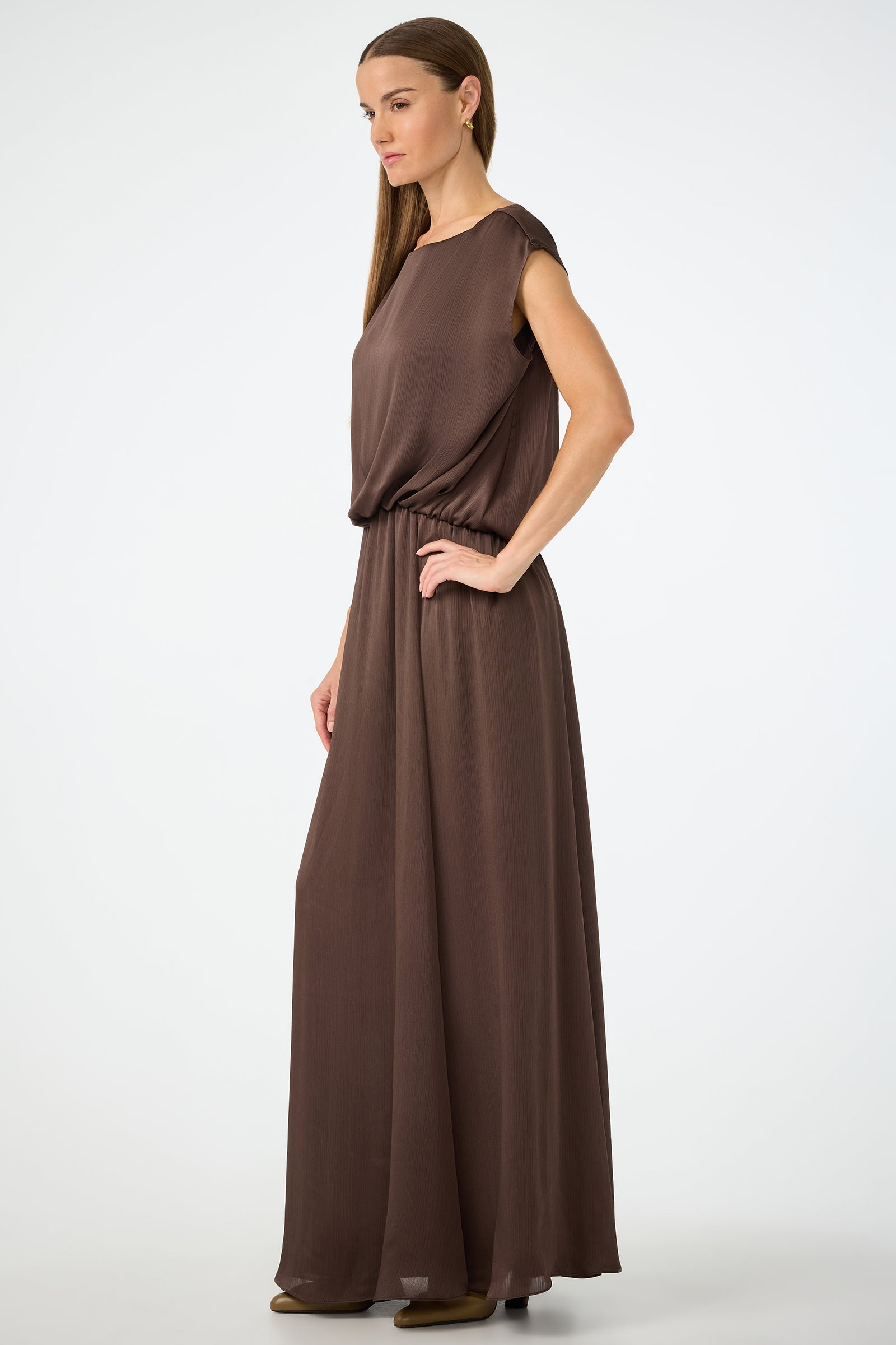 A woman with long straight hair, in profile against a white background, wears the Vantage 2 Maxi Dress—a sleeveless, floor-length brown dress with a cinched waist.