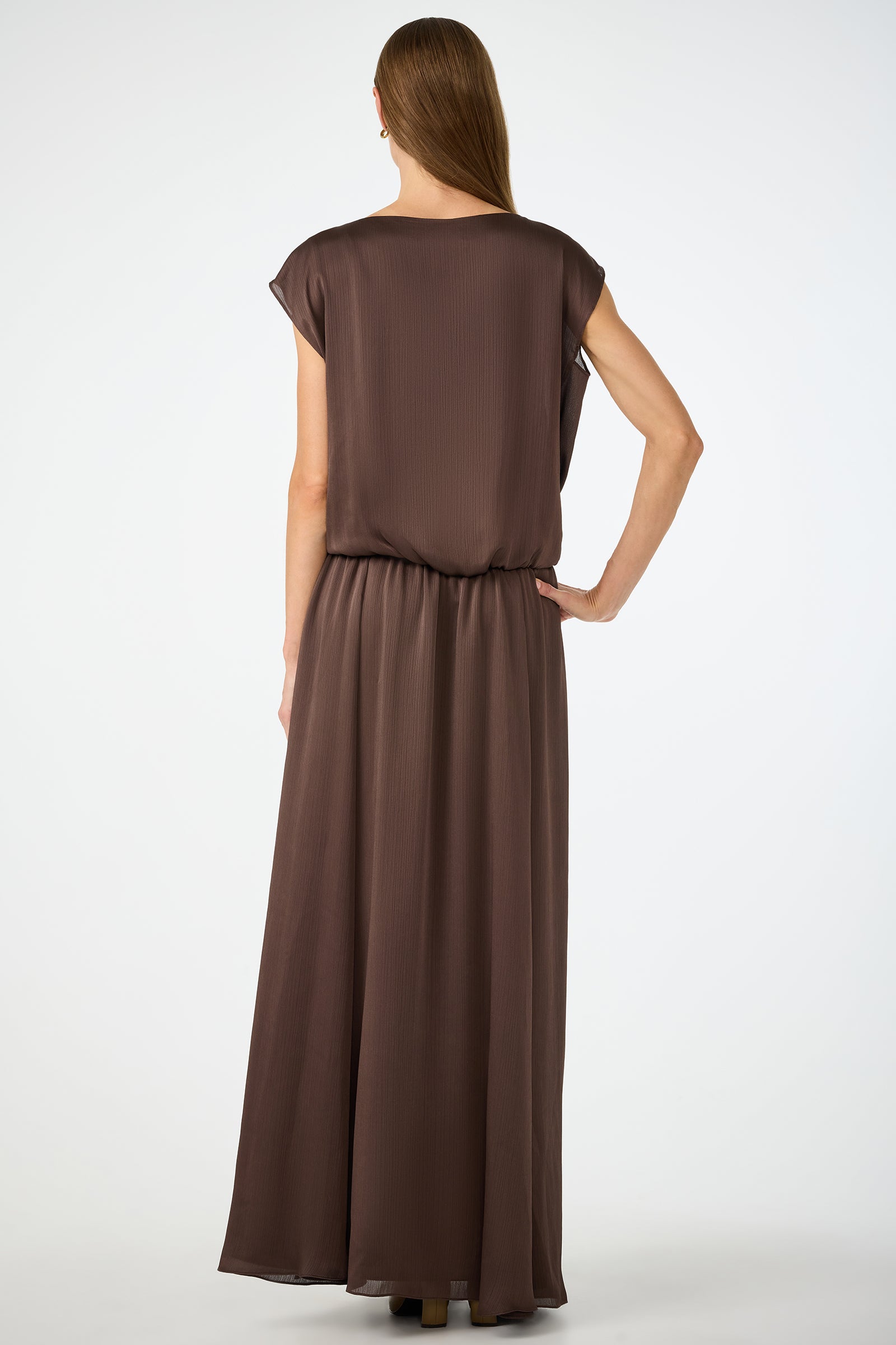 A woman with long, straight hair stands with her back to the camera, wearing the Vantage 2 Maxi Dress, a loose, short-sleeved, floor-length brown dress. The background is plain white.