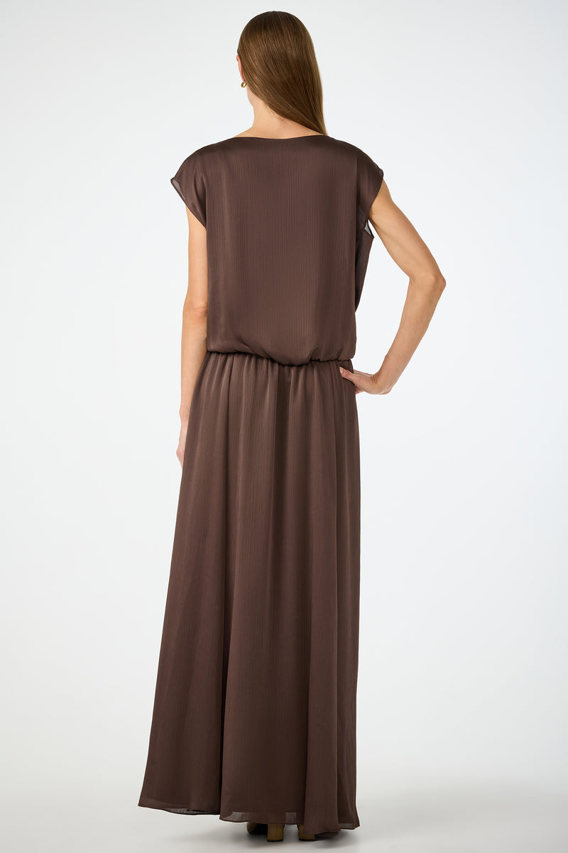 A woman with long, straight hair stands with her back to the camera, wearing the Vantage 2 Maxi Dress, a loose, short-sleeved, floor-length brown dress. The background is plain white.