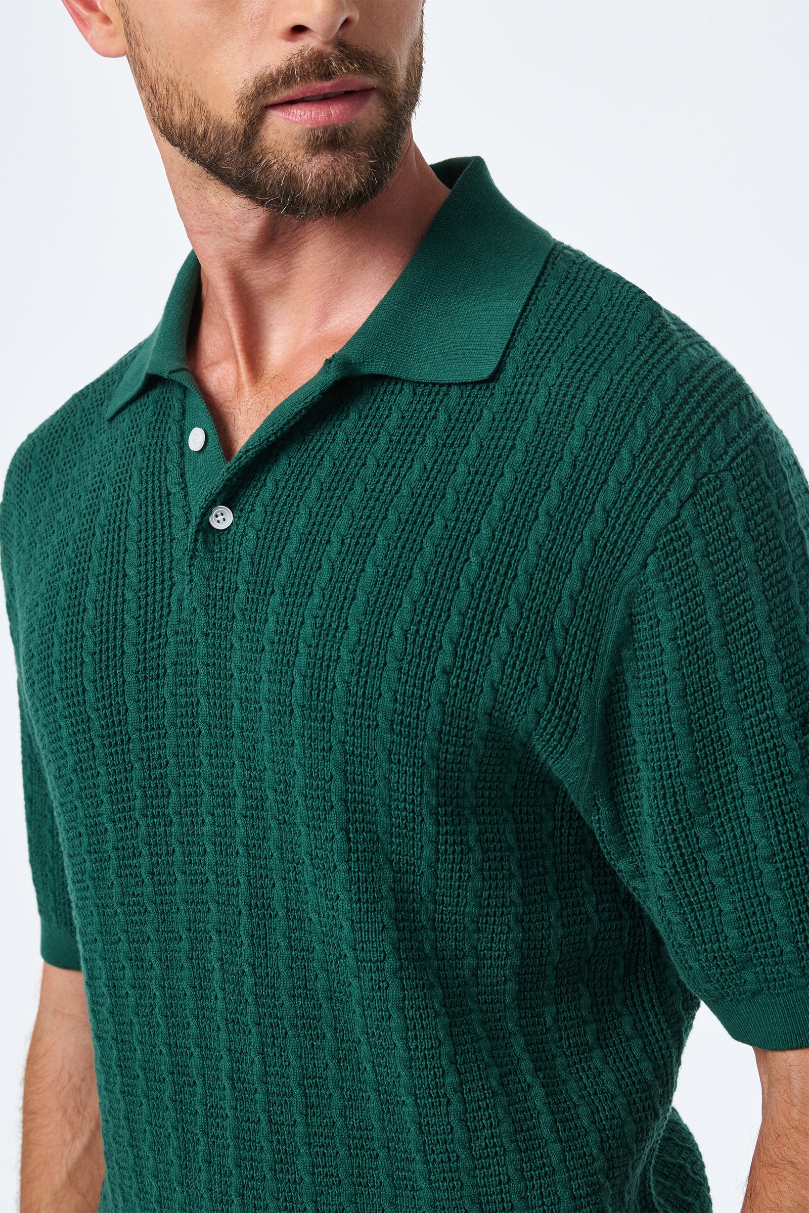 A man with a trimmed beard wears the Asher Ss Polo, a textured dark green short-sleeve knit shirt with a collar and two buttons, looking slightly to the side against a plain white background.