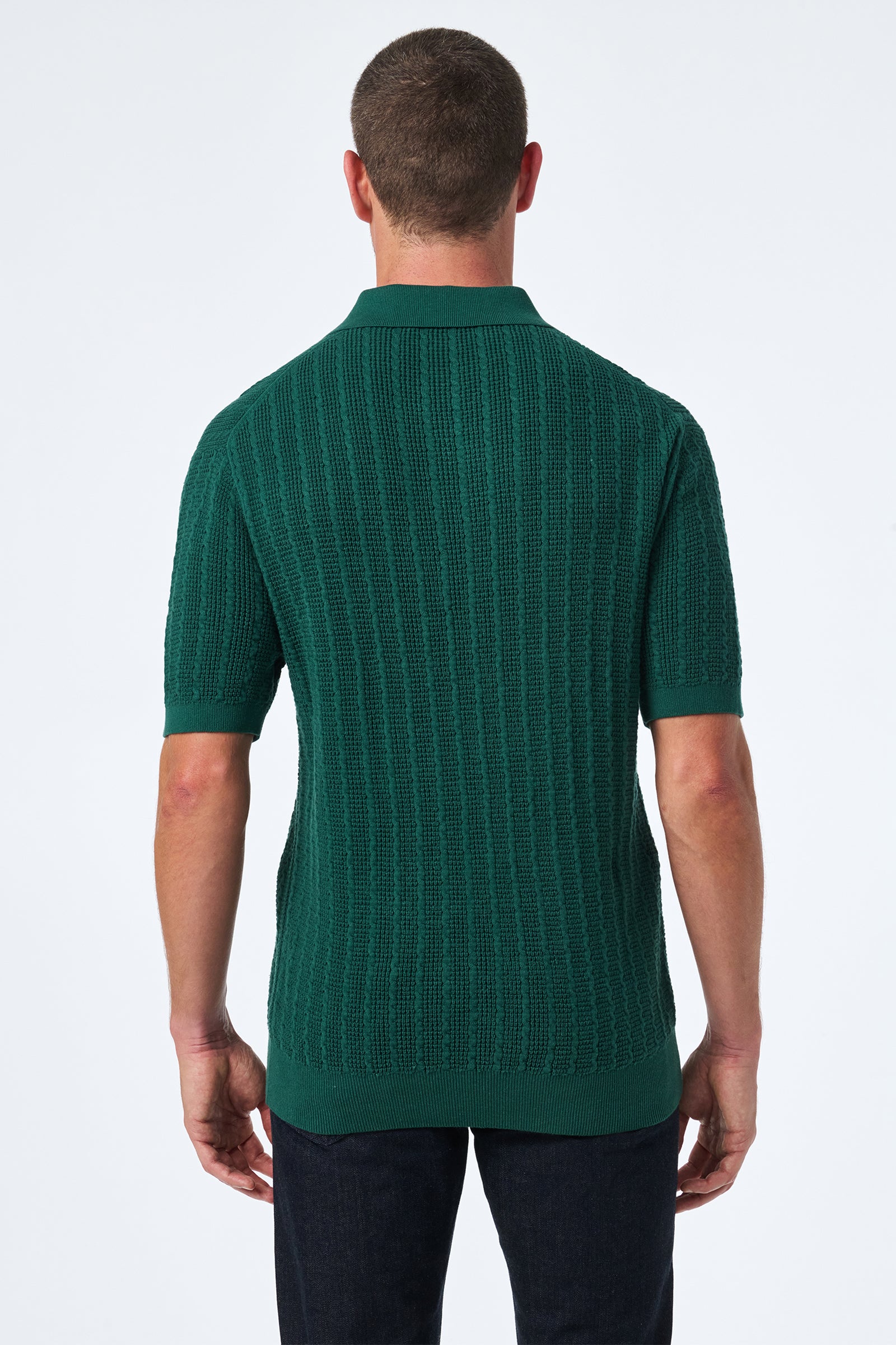 A man stands facing away, wearing the Asher Ss Polo—green, short-sleeved, knit with a ribbed texture—and dark pants against a plain light background.