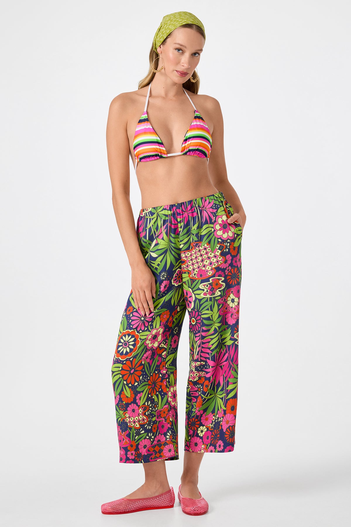 A woman poses in a colorful striped bikini top, Estella 2 Cropped Pants - Fumi Floral, a green patterned headscarf, and pink shoes against a plain white background.