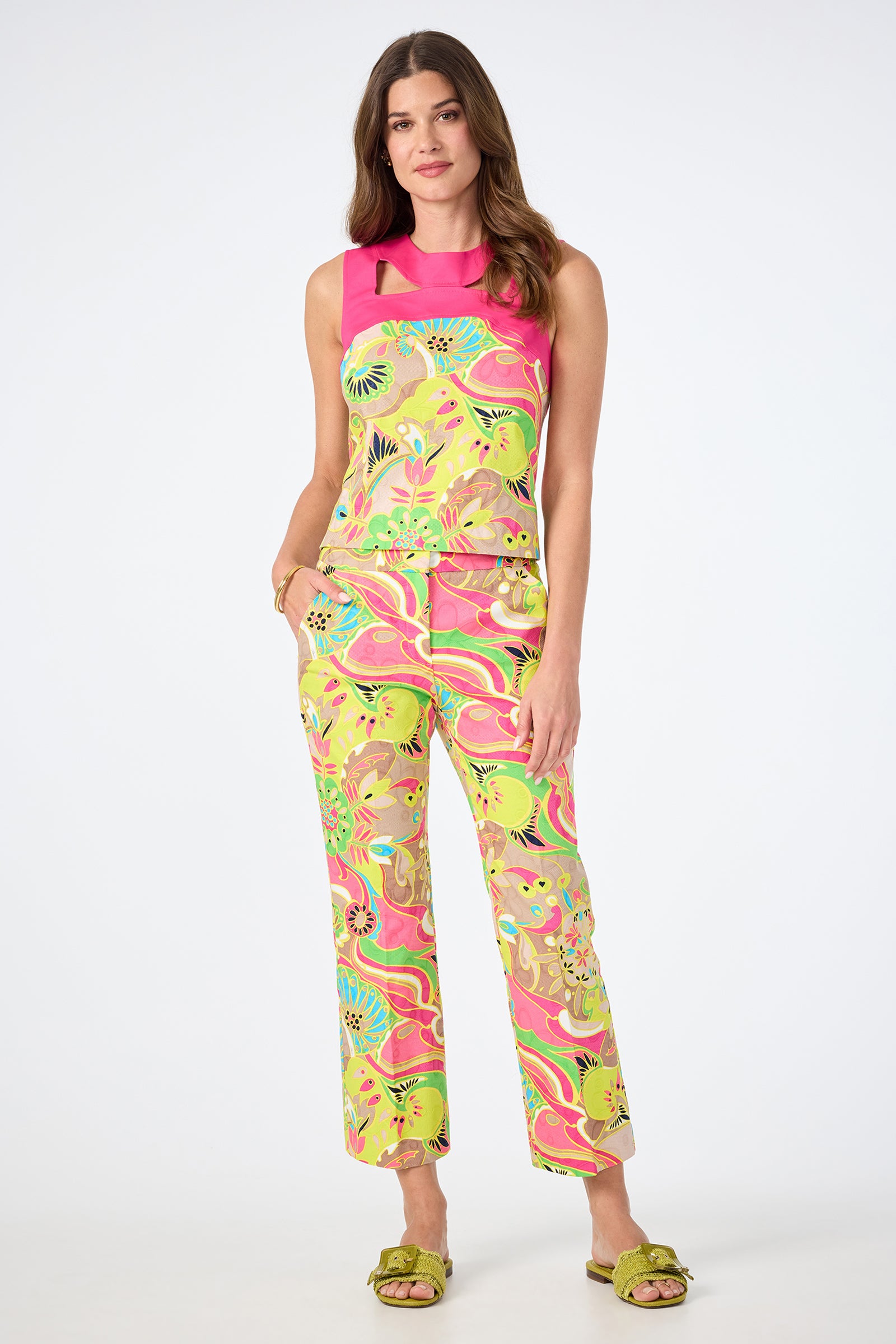 A woman with long, wavy hair stands against a plain background, wearing the Lulu Cropped Pant - Venus and a matching sleeveless top featuring vibrant pink, yellow, green, and brown floral and abstract patterns. She pairs the look with green slide sandals.