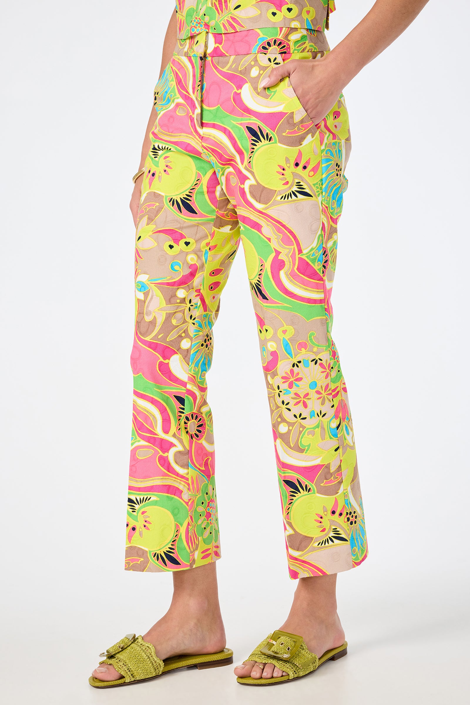 A person wearing the Lulu Cropped Pant - Venus, featuring colorful swirls and floral designs in yellow, pink, green, and beige, paired with green textured slide sandals, stands against a plain white background.