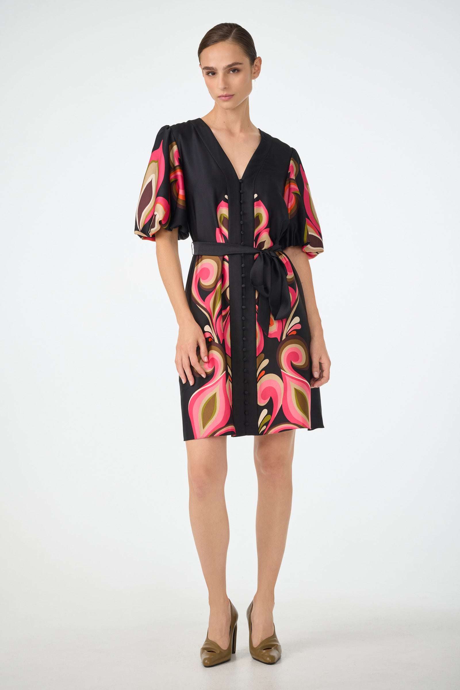 A woman stands in a studio wearing the Certonia Puff Sleeve Dress - Sanctuary, featuring short puff sleeves, a belted waist, and Sanctuarys signature colors, styled with bronze high heels.