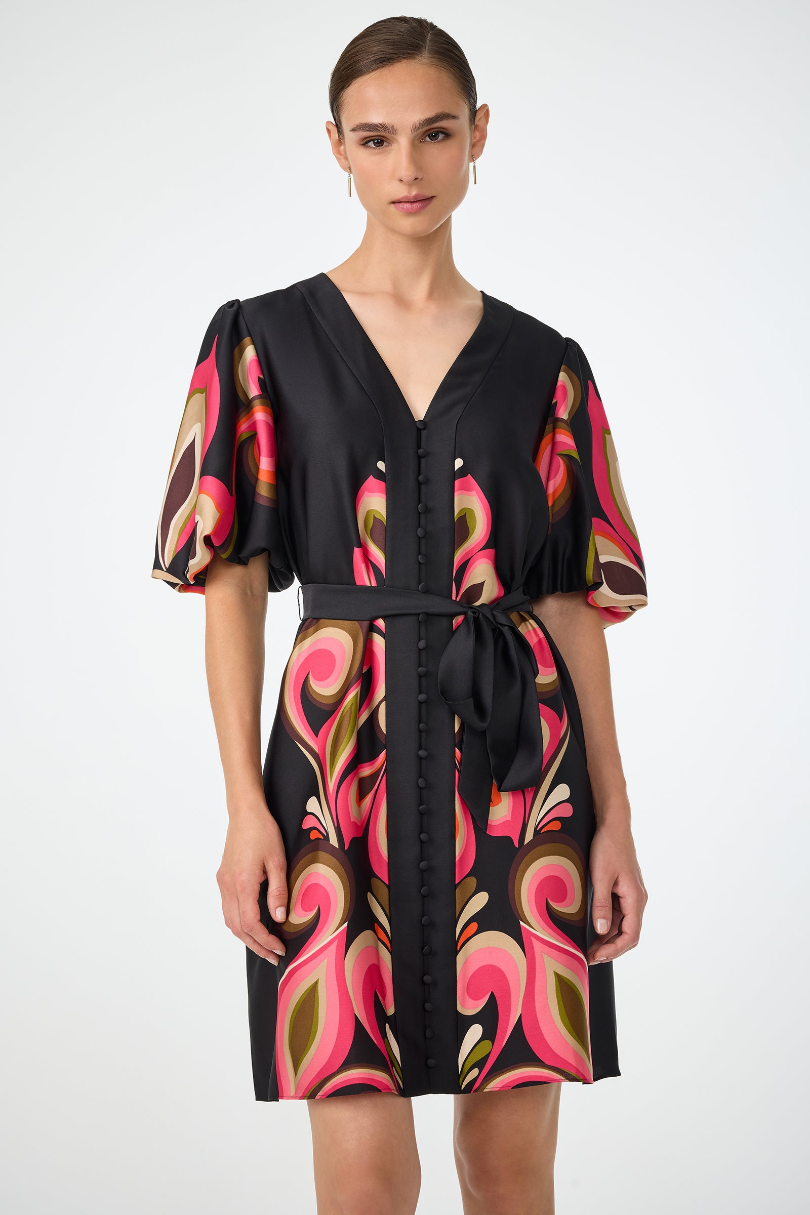 A woman models the Certonia Puff Sleeve Dress - Sanctuary, featuring a black base with swirling pink, orange, and olive green patterns, puffed short sleeves, a V-neck, front buttons, and a matching waist tie against a white background.