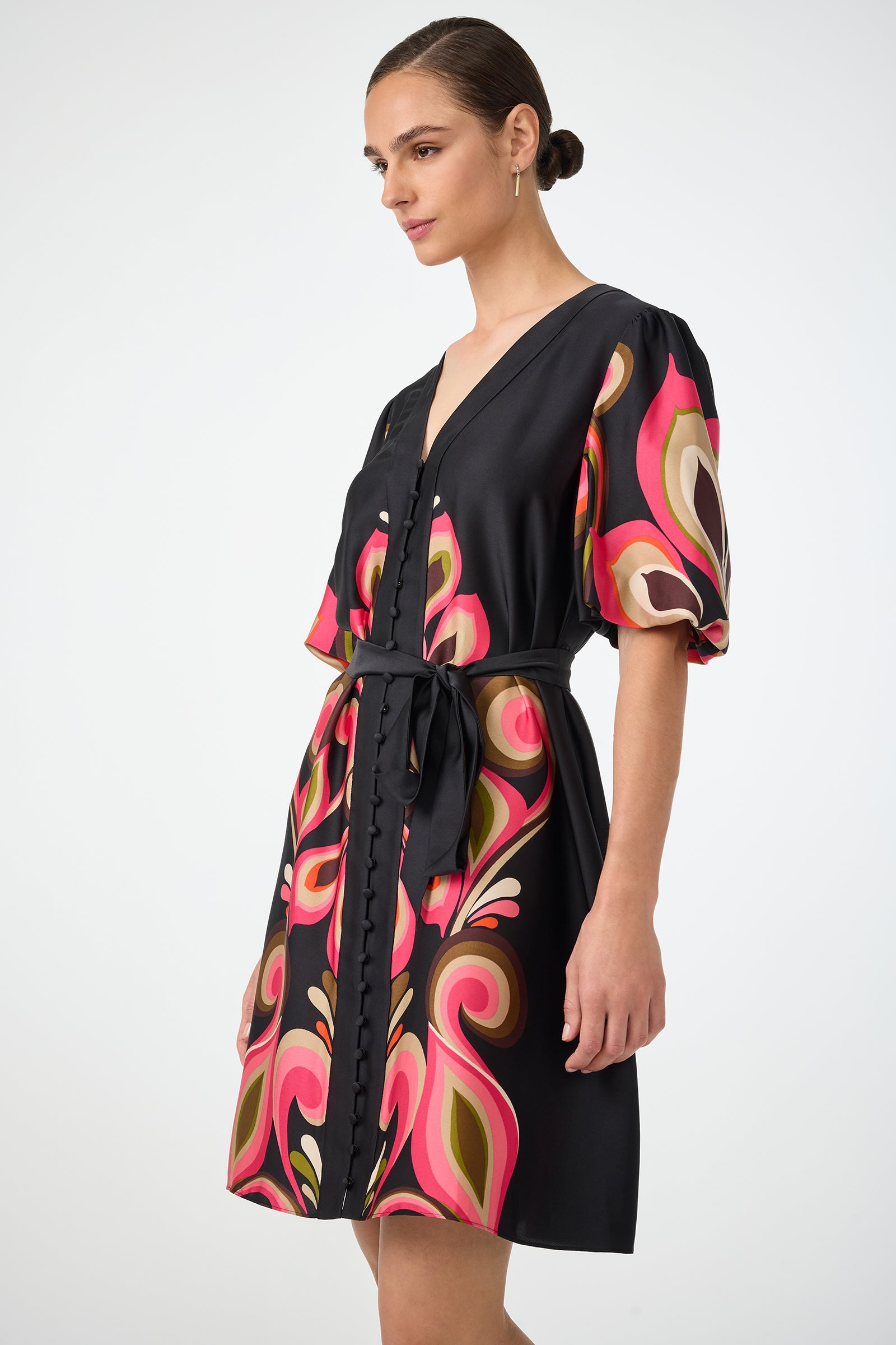 A woman wearing the Certonia Puff Sleeve Dress by Sanctuary, featuring a black base with colorful abstract pink, red, and olive green prints, a V-neckline with button details, a tied waist belt, and her hair styled in a sleek bun.