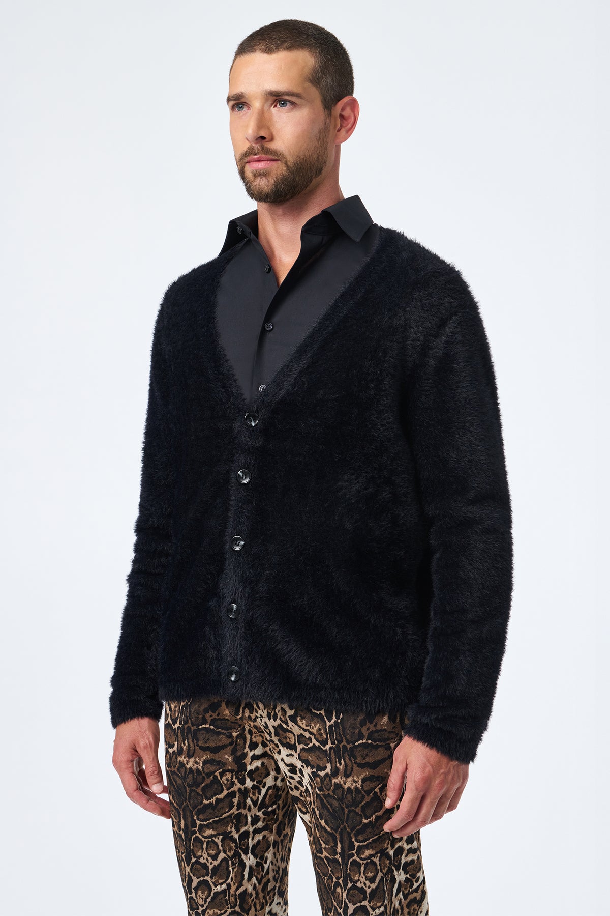 A man with short hair and a beard wears the Scott Fuzzy Cardigan in black over a black shirt and leopard print pants, standing against a plain white background.