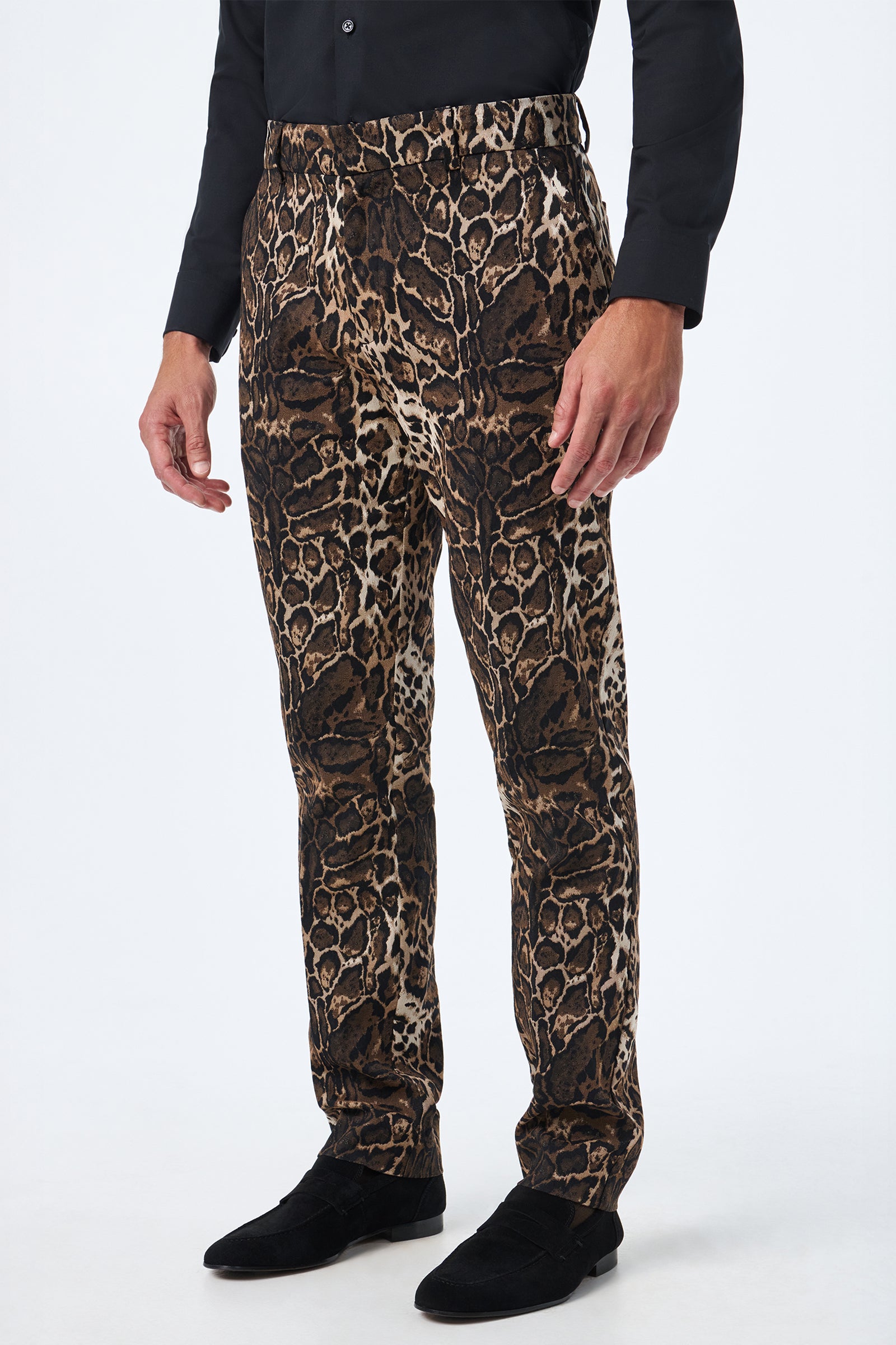 A person wearing a black long-sleeve shirt, black shoes, and the Clyde Slim Trouser - Lompoul Leopard stands against a plain white background. The trousers have a bold leopard print pattern.