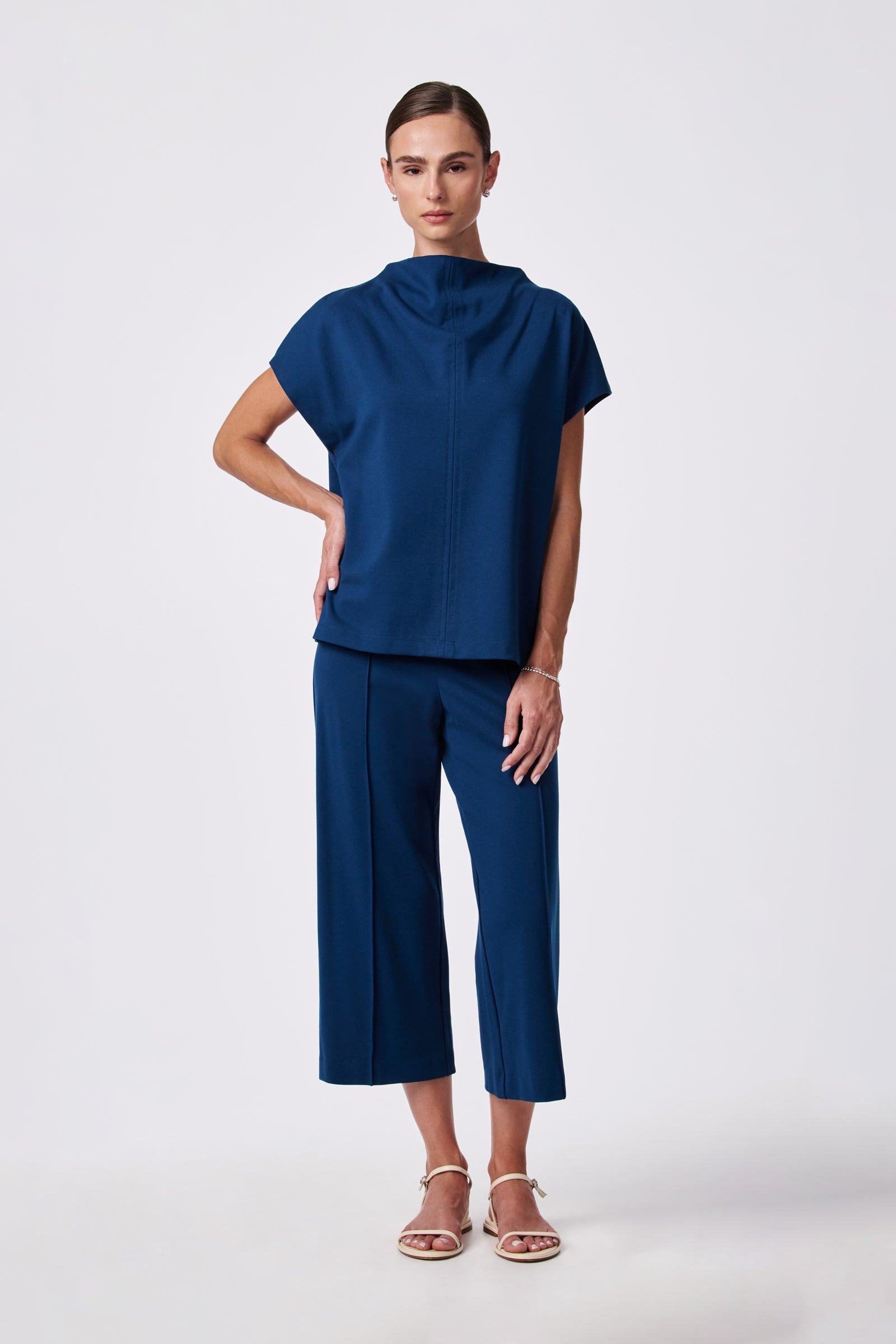 A person stands against a plain background wearing the Pavilion Top, a loose-fitting, short-sleeved blue top with a high neckline, paired with matching blue cropped pants and white sandals.