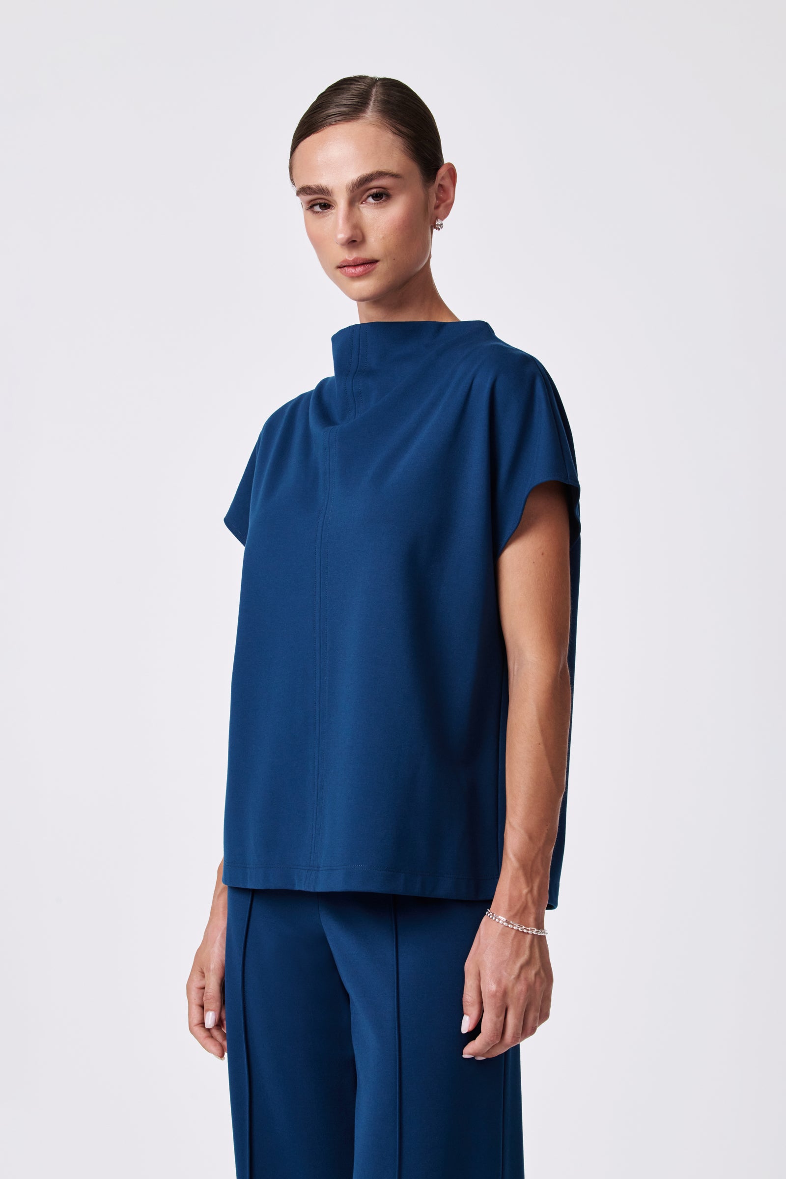 A woman with dark hair in a low bun wears the Pavilion Top, a loose-fitting, short-sleeved, high-neck blue top, paired with matching wide-leg pants against a plain light background.