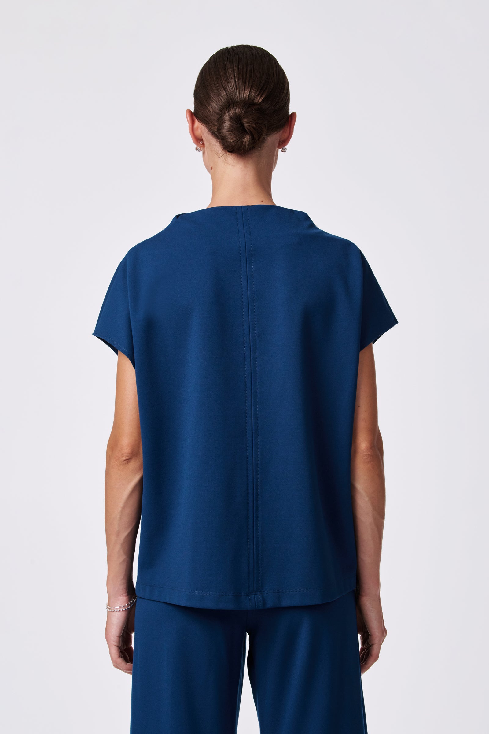 A person with brown hair in a neat bun is seen from behind, wearing the loose-fitting, short-sleeved Pavilion Top in dark blue with matching pants against a plain light background.