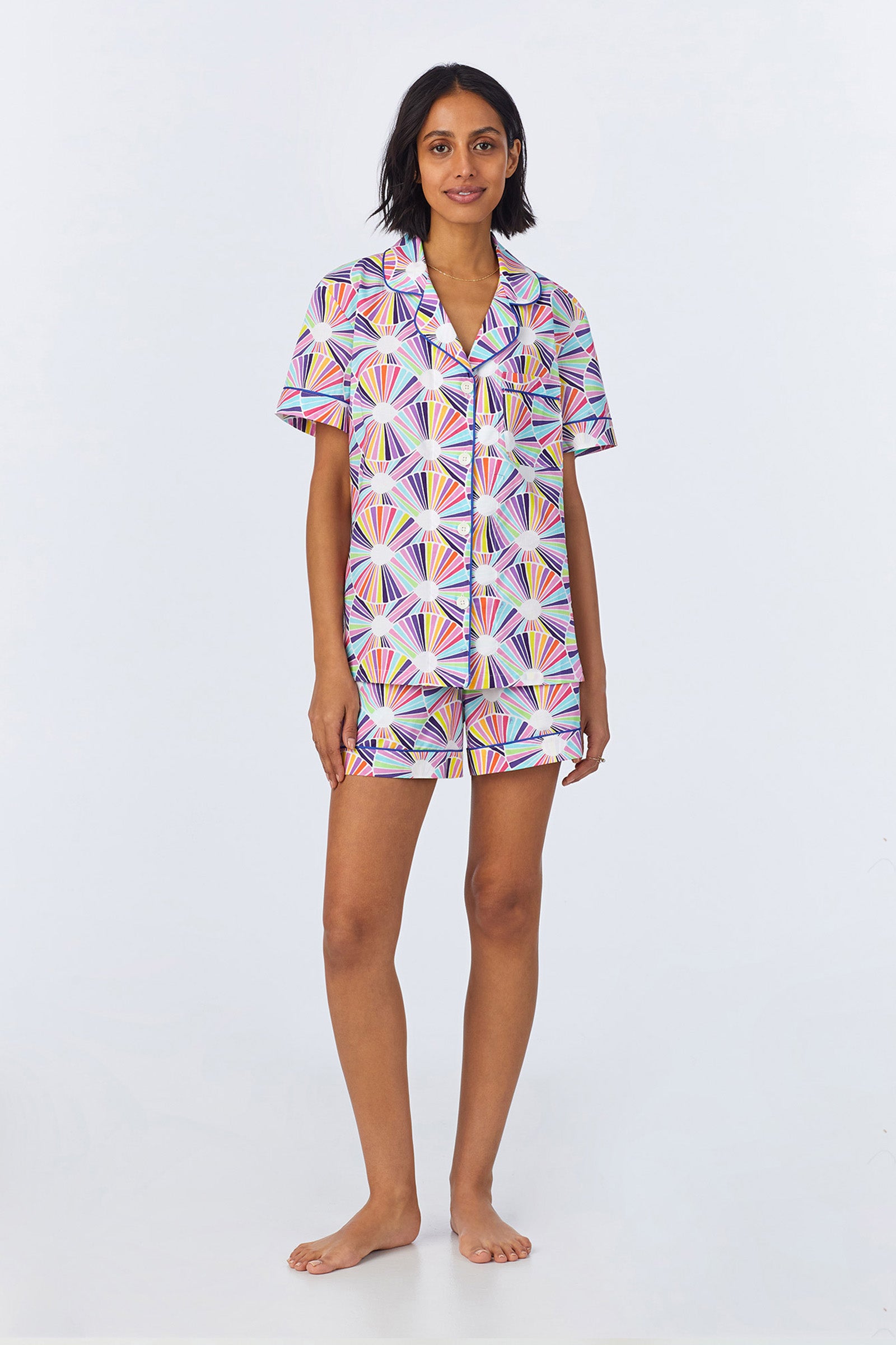 A woman stands barefoot against a white background wearing the Shorty PJ Set - Fanfare Poplin in Multi, featuring a matching button-up short-sleeve shirt and shorts with a colorful geometric pattern.