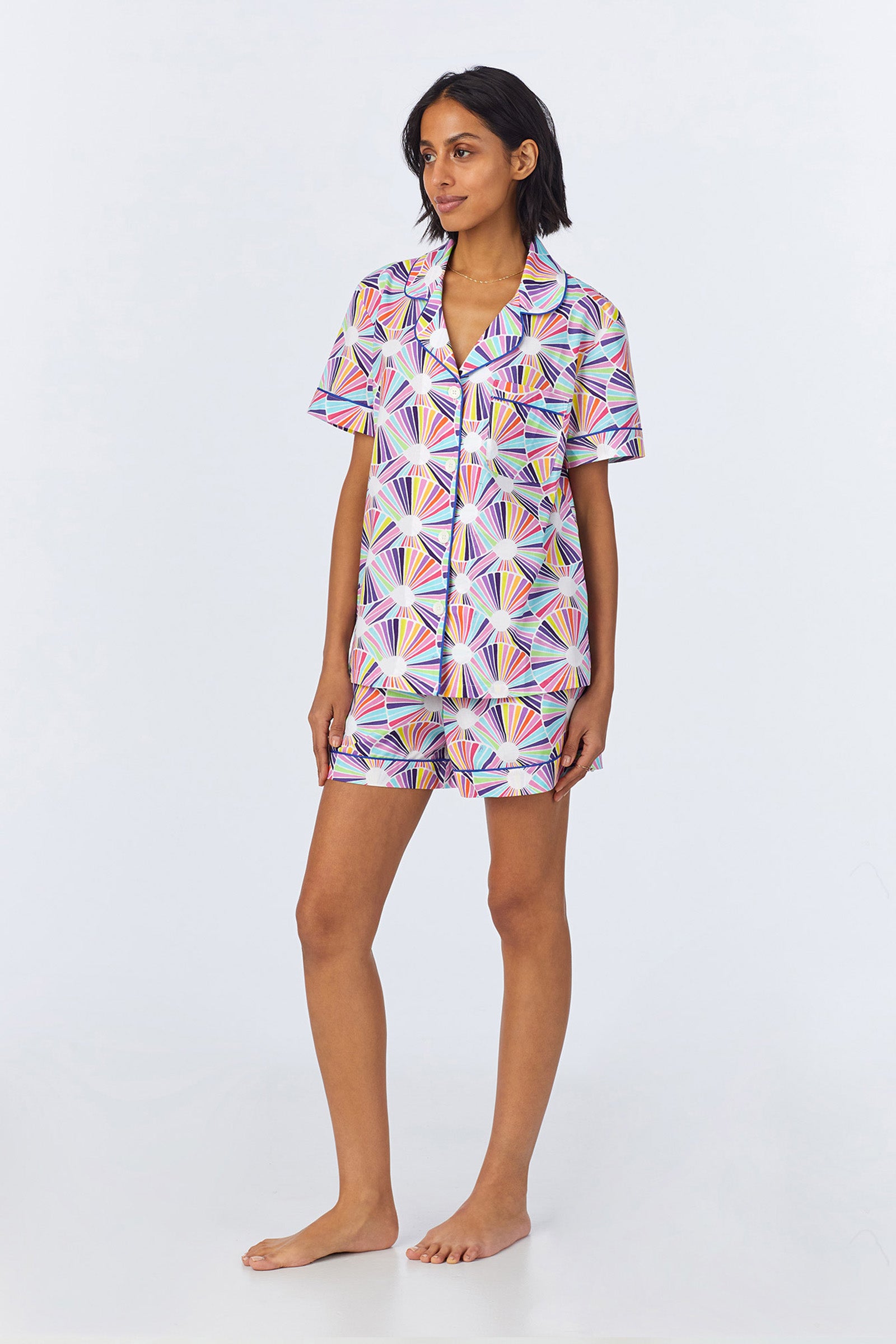 A woman stands barefoot in the Shorty PJ Set - Fanfare Poplin, a colorful matching short-sleeve set with a geometric fan pattern. The button-up collared shirt and shorts both showcase the same vibrant design.